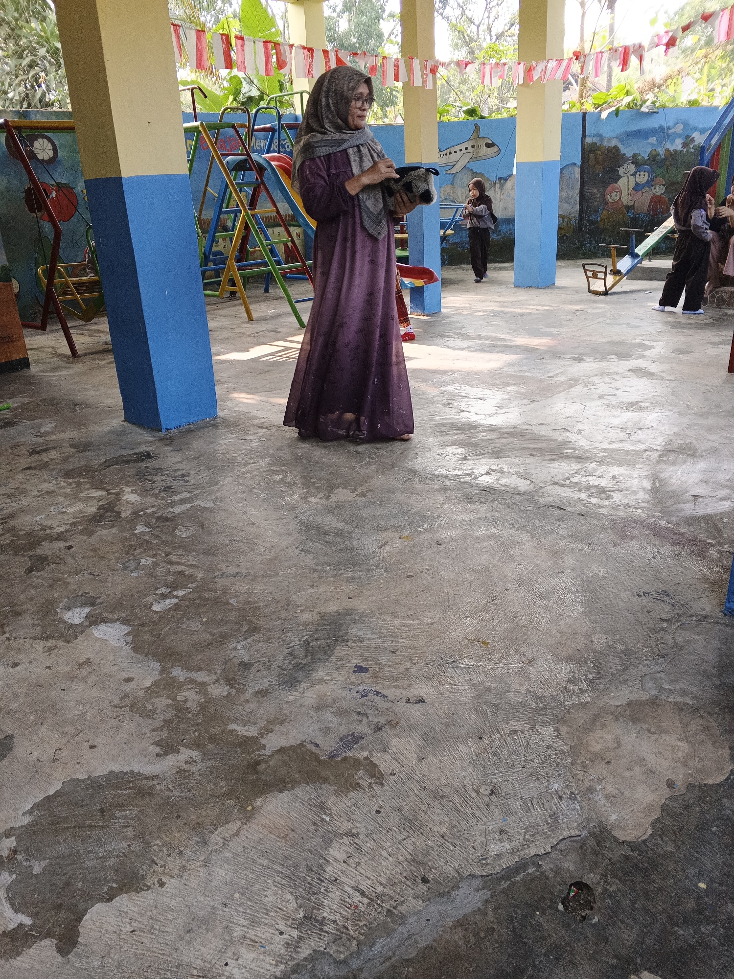 In Warungkiara, Indonesia, an outdoor playground area features a woman in a purple hijab and dress holding a stuffed animal. She stands near a blue and yellow pillar and a colorful playground structure. Behind her, a mural adorns the wall, depicting children playing and an airplane. Several children are also present, some in hijab and uniform, engaged in activities like using a seesaw. Red and white Indonesian flags are strung overhead, suggesting a national celebration. The ground is a concrete surface with visible wear and tear.