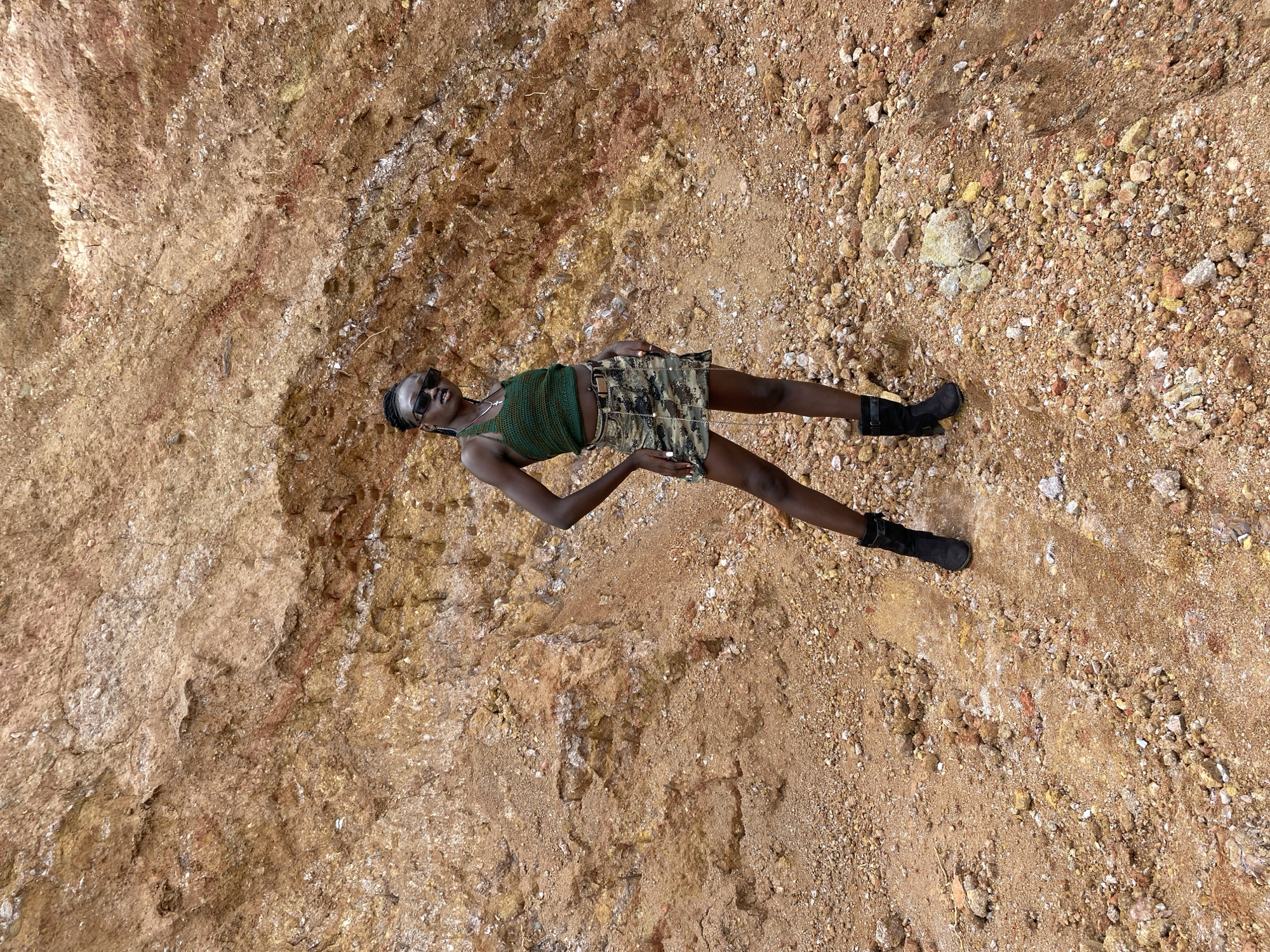A young Black woman wearing a green crochet tank top, a camouflage skirt, black boots, and sunglasses stands posing in front of a reddish-brown, rocky cliff face in Bar Jirgi Summa, Nigeria. The ground around her is covered in loose rocks and dirt. The woman is positioned in the center of the frame, facing slightly to her left. Her left hand is on her hip, and her right arm is bent, with her hand resting on her thigh. The cliff face behind her shows layers of different colored soil and rocks, with some lighter-colored veins running through it. The lighting suggests it is daytime, with no harsh shadows indicating direct sunlight.