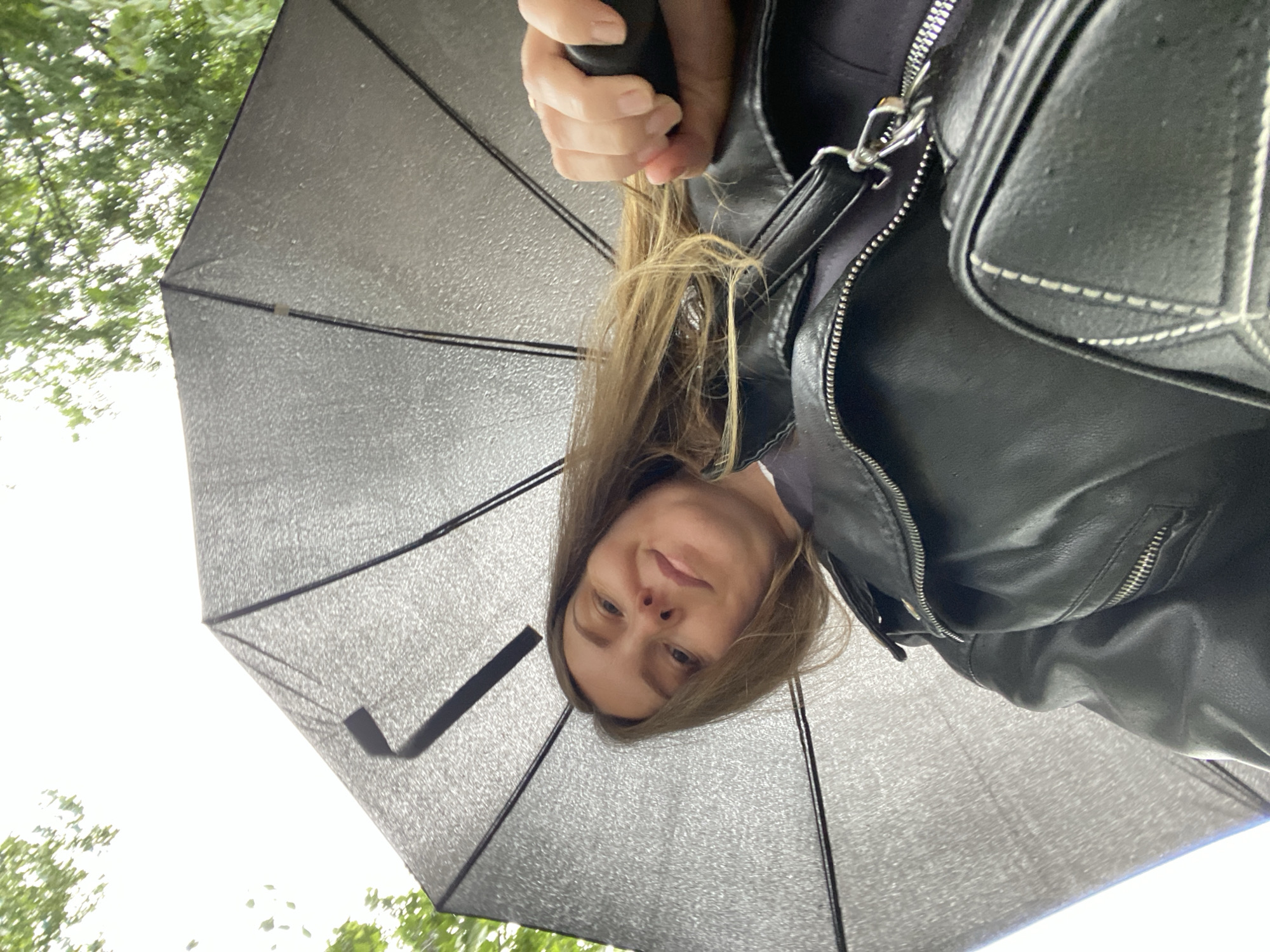 A woman stands under an open black umbrella, with raindrops visible on its surface. She wears a black leather jacket. Some trees and sky are visible in the background. The location is unspecified, and there is no verifiable connection to Noviy, Russia.