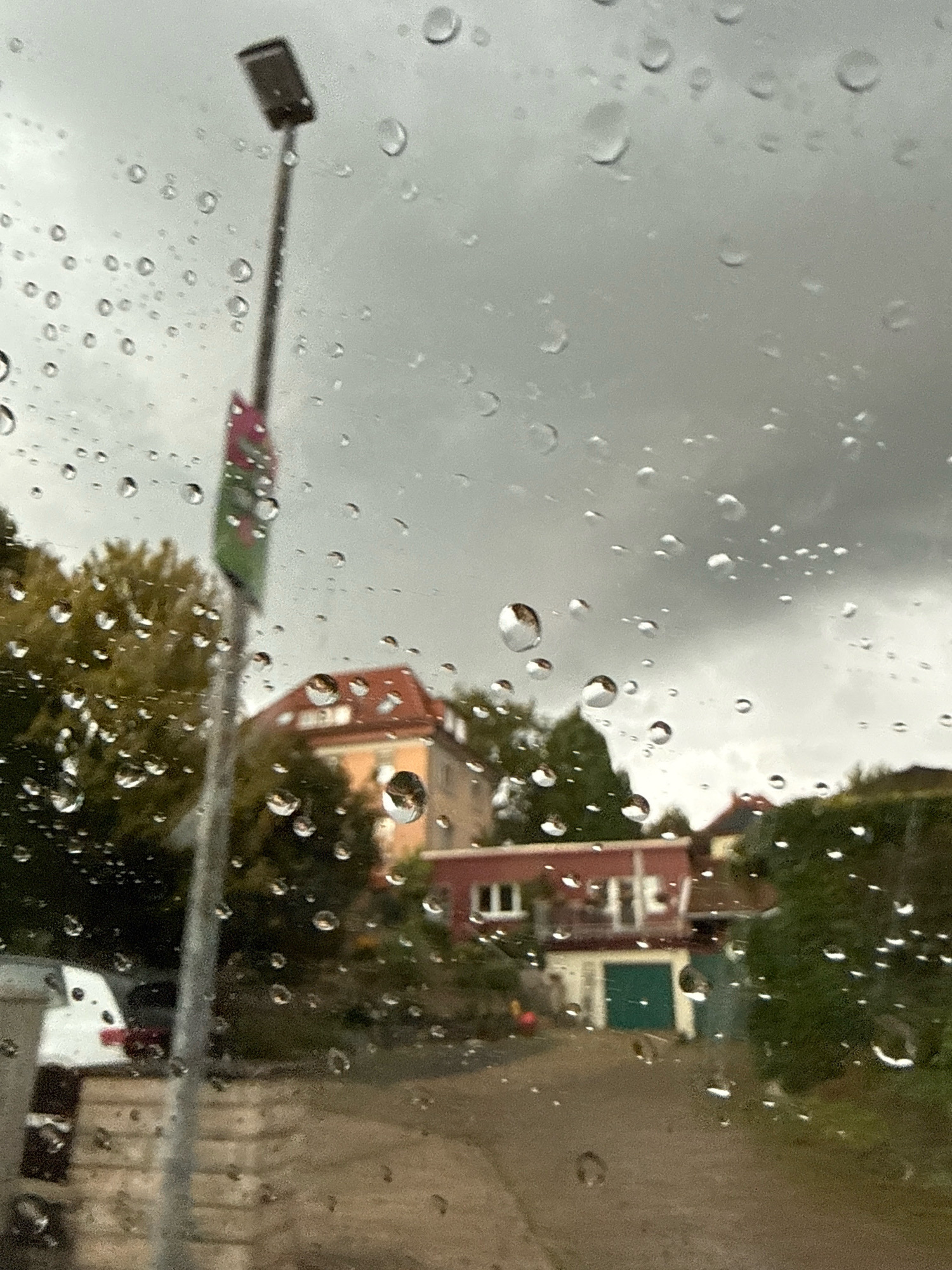 A medium-shot image taken through a window with raindrops shows a street scene in the town of Bad Sachsa, Germany. A tall, thin lamp post with a rectangular light fixture stands in the foreground to the left, adorned with a partially visible green and pink banner. Beyond the lamp post, a two-story building with a red-tiled roof is visible, featuring multiple windows and a balcony. Adjacent to it is a smaller building with a green garage door. Trees with autumn foliage line the street, and a white car is parked on the left side. The sky is overcast and gray, indicating rainy weather. The ground appears to be a paved road or driveway. The scene is out of focus due to the raindrops on the windowpane.