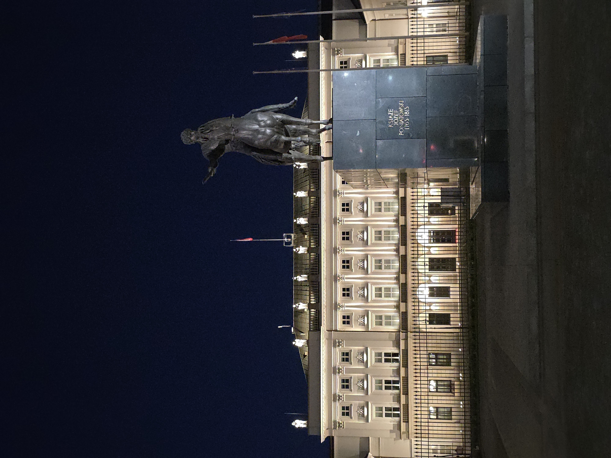 The image captures a nighttime scene in Warsaw, Poland. The prominent feature is an equestrian statue, likely depicting Prince Józef Poniatowski, standing atop a tall, rectangular plinth with an inscription. Behind the statue is a large, illuminated building, possibly a palace or government building, with symmetrical windows and architectural details. The foreground includes a wrought-iron fence separating the viewer from the building. A dark blue, almost black, sky dominates the left portion of the image, indicating the late hour. Flags are visible atop the building, adding to the formality of the scene. The overall impression is one of historical grandeur and urban elegance.