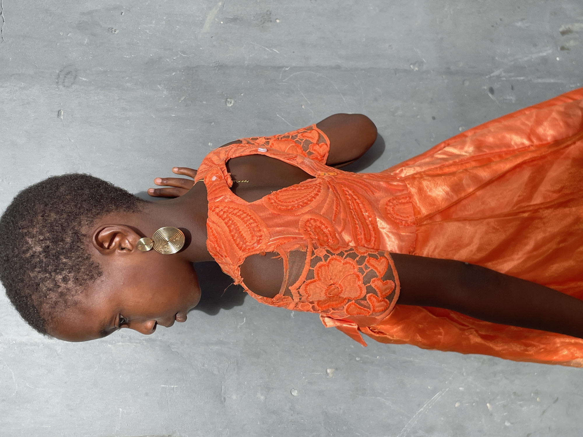 A woman wearing an orange dress lies on a grey, matte surface. The dress is knee-length and appears to be made of both a lace material and a shiny fabric. The woman's head is positioned to the left, and she wears gold, round earrings. The background is a flat, grey surface, possibly a concrete floor. The setting suggests an indoor or enclosed space. The image does not contain enough information to determine the exact location or any relation to the city of Badagry, Nigeria.