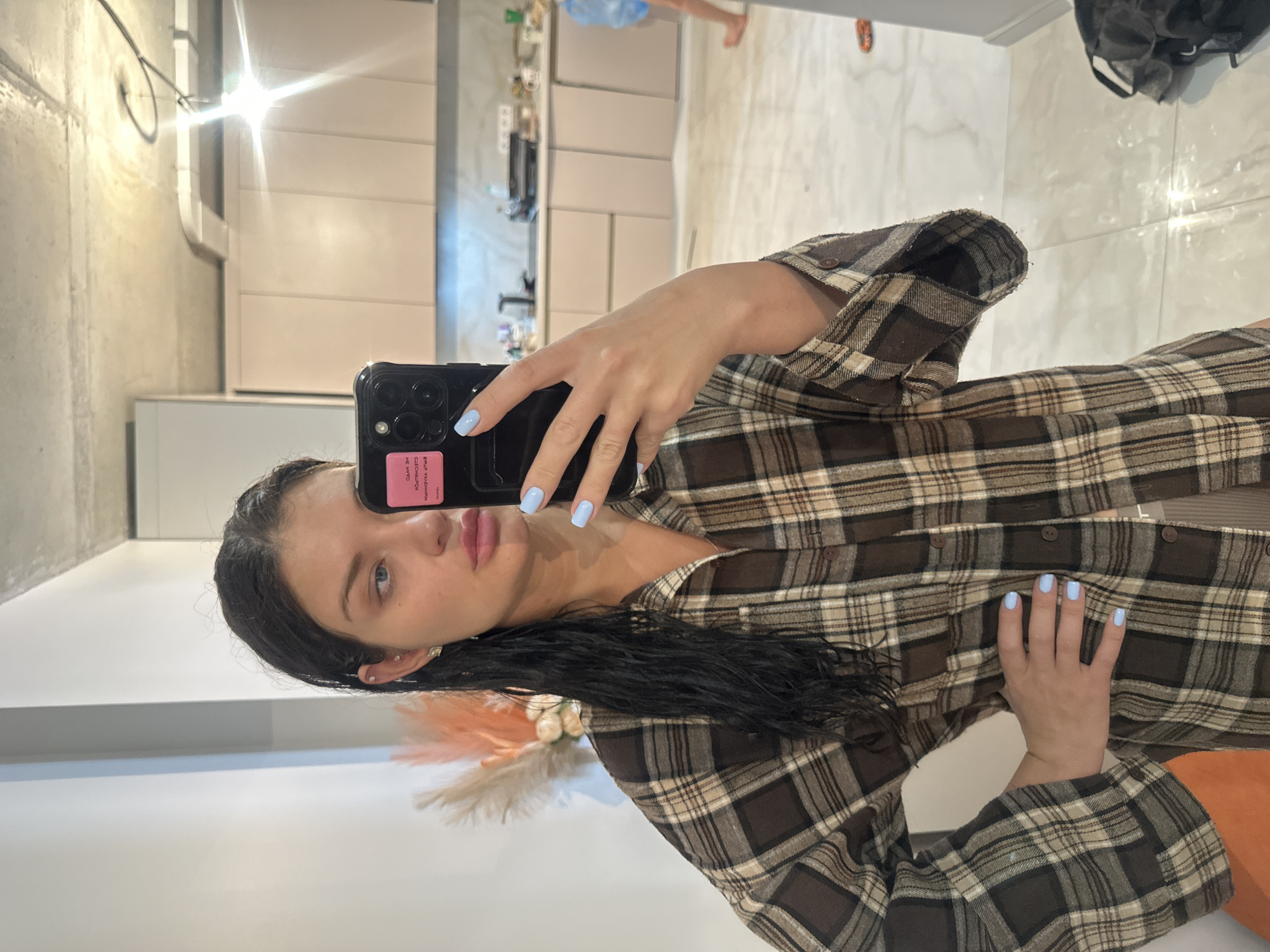 A woman with dark hair and blue eyes takes a selfie in front of a mirror. She is wearing a brown and tan plaid shirt. Her nails are painted light blue. The mirror reflects a kitchen with light brown cabinets and a marble floor. In the reflection, another person's legs are visible. A black bag is on the floor. There is no evidence that this image was taken in Moscow, Russia.