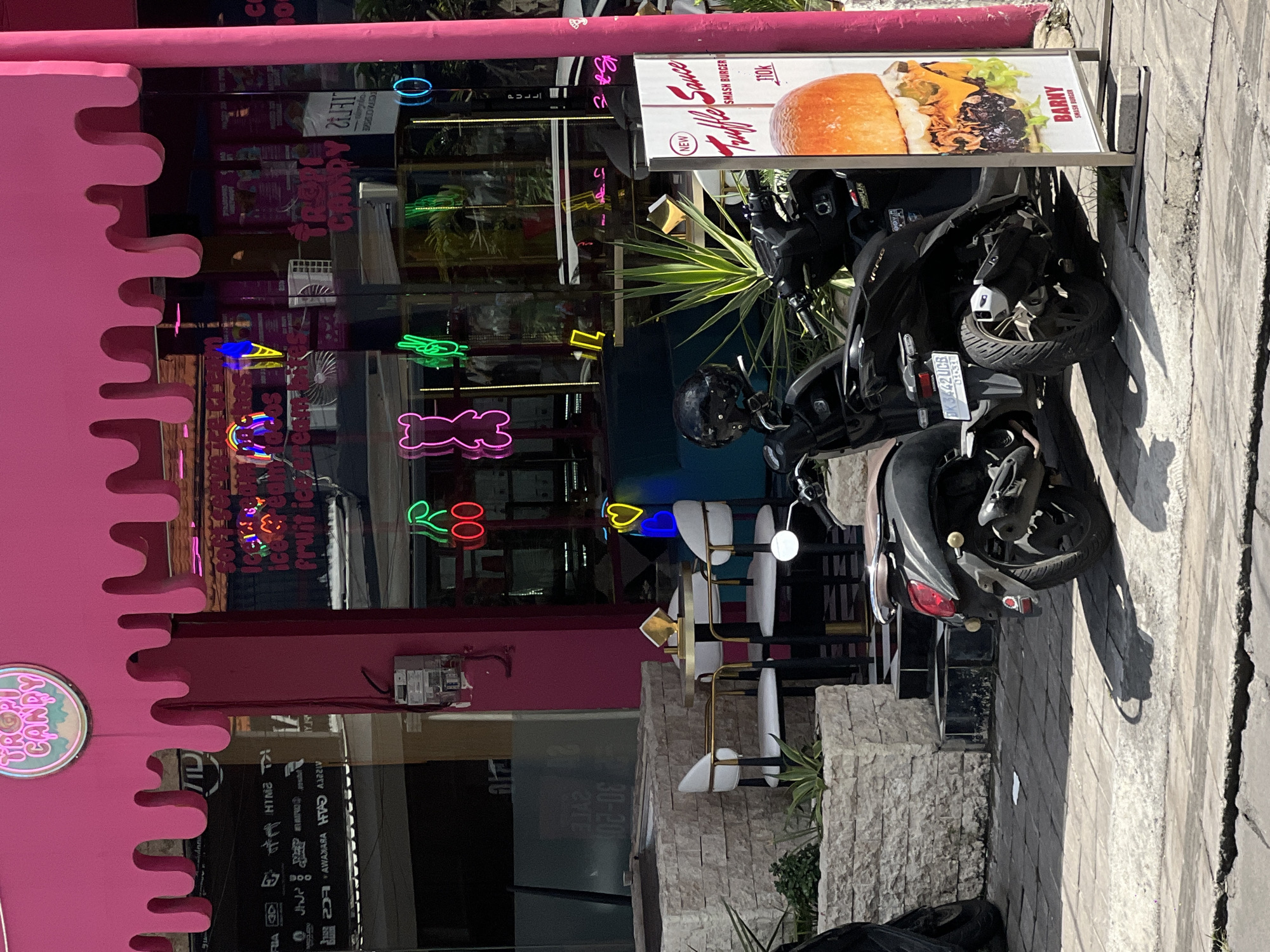 This vibrant image captures a lively street scene in North Kuta, Indonesia, showcasing a unique dessert shop with parked motorcycles. The central subject is a commercial building with a striking bright pink facade, featuring a playful, wavy "dripping" design along its edges. A circular sign on the pink exterior reads "TROPI CANDY."
Behind large glass windows, numerous colorful neon signs illuminate the interior, depicting whimsical shapes such as a bear, a cherry, an ice cream cone, and a rainbow. Visible text on these signs and reflections includes "TROPI CANDY," "soft cream," and "fruit ice cream."
Outside the shop, an inviting outdoor seating area is furnished with modern white and gold bar stools and small tables. Adjacent to this, a tall vertical sign advertises a "NEW Truffle Sauce SMASH BURGER" from "BARNY SMASH BURGER" for "110K." Several black motorcycles and scooters are parked on the paved sidewalk in front of the establishment, one displaying the license plate "DK 3442 UCB."
The scene is bathed in bright sunlight, casting long, distinct shadows from the motorcycles and the vertical sign, indicating it is daytime, likely mid-morning or afternoon, under clear skies. The overall atmosphere is cheerful, vibrant, and inviting, characteristic of a popular tourist destination. Other discernible text includes a reflected "PULLL" on the window and "SOMPLAK" on the burger sign. No people are visible in the frame.