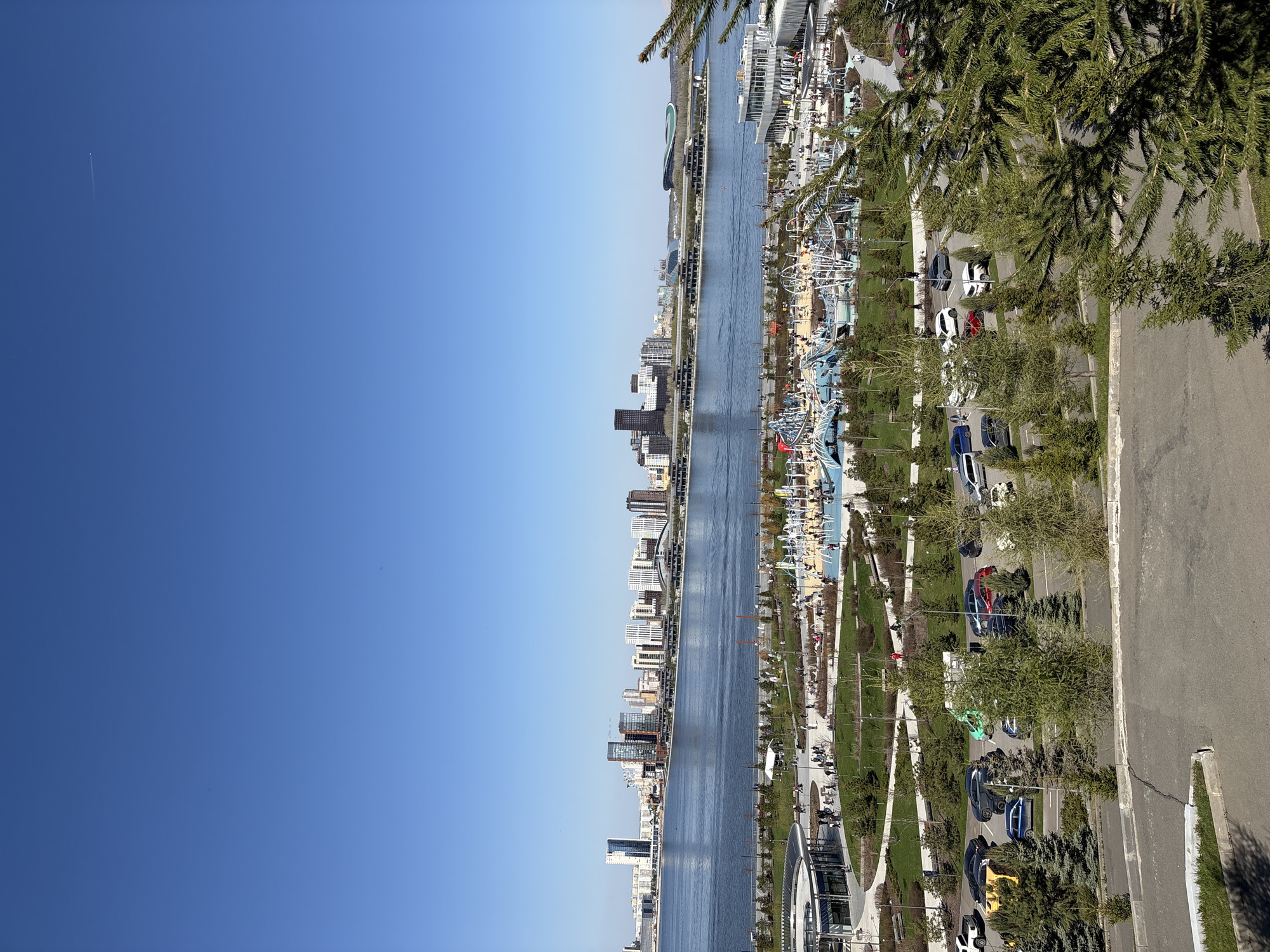 The image shows an aerial view of Kazan, Russia. A wide river runs through the center of the frame, with buildings lining its banks. On one side is a modern city with skyscrapers and residential buildings. The opposite side features a park with trees, walking paths, and a variety of attractions, including what appears to be a Ferris wheel and other amusement park rides. A road runs parallel to the park, with parked cars visible alongside the trees. The sky is clear and blue. Trees are visible on the right edge of the frame, adding a natural element to the urban landscape.