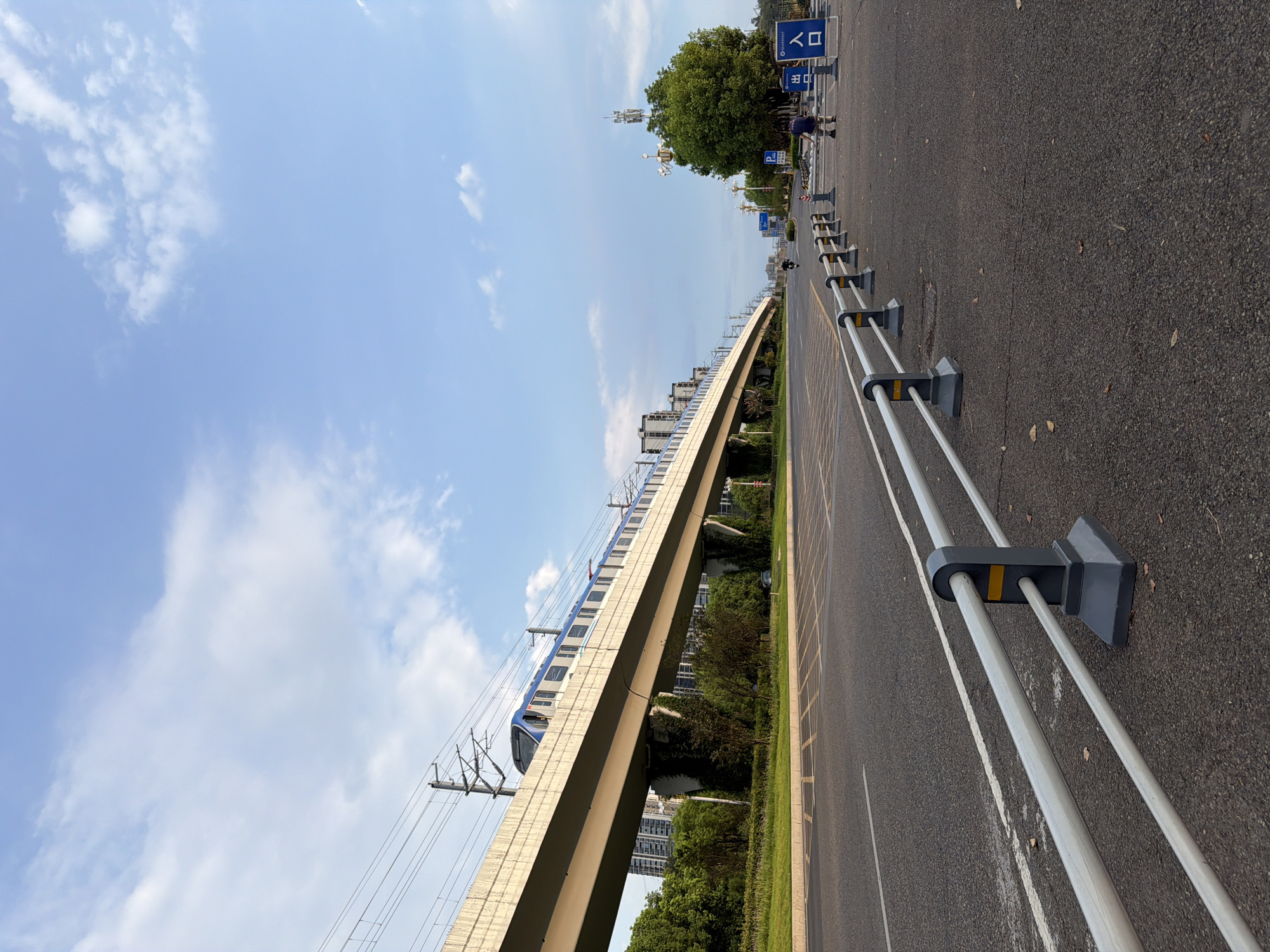 In Jiangjin, China, a metro train travels on an elevated track over a wide asphalt road. The road has lane markings and a metal barrier along its side. A person is walking on the sidewalk to the right of the road, near a green tree and a blue sign that reads "入口," meaning "entrance." The sky is blue with scattered clouds, and the scene is bathed in natural light, suggesting it's daytime. Buildings are visible in the background, indicating an urban setting.