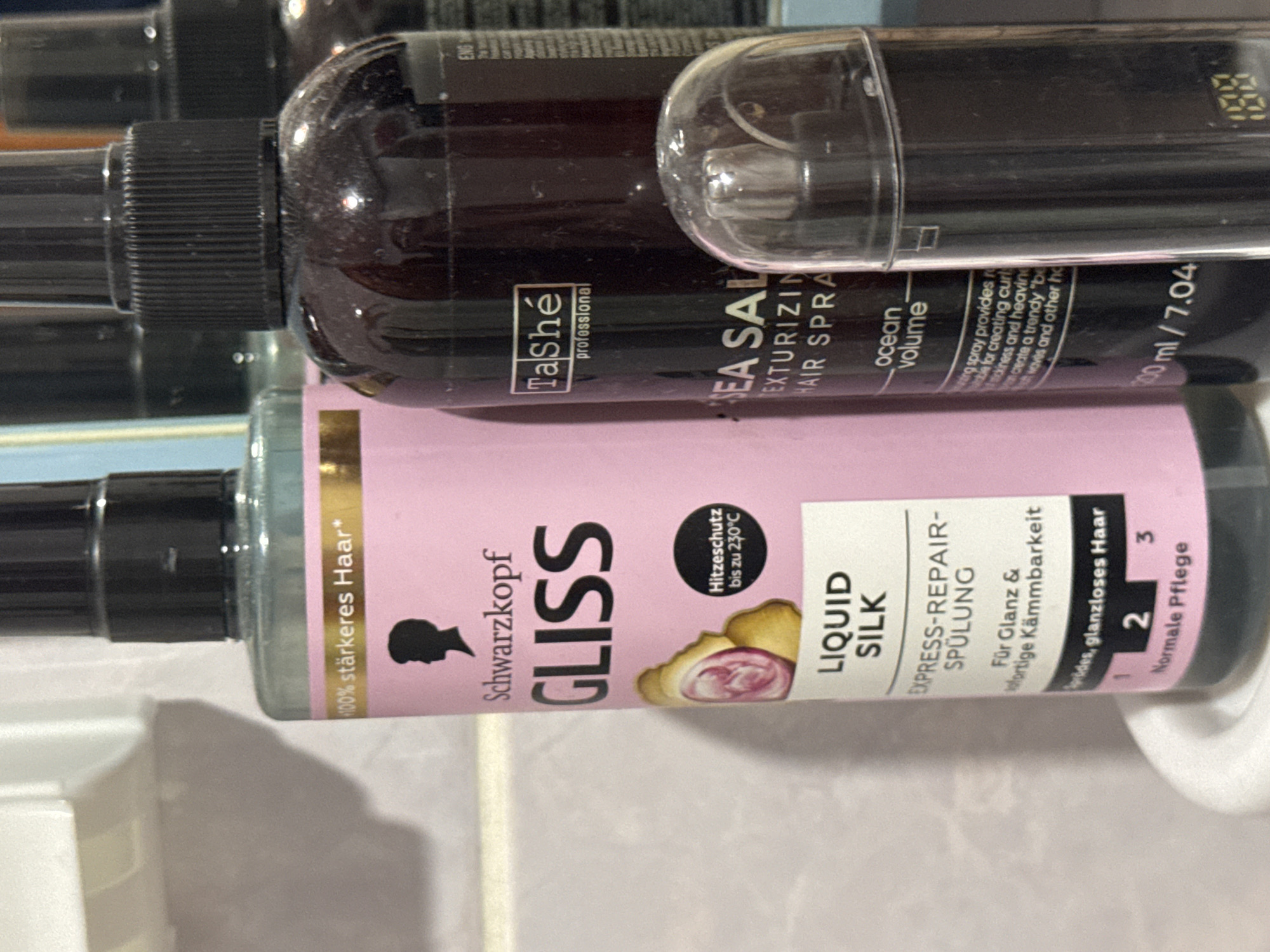 The image shows a close-up, eye-level view of three hair products lined up on a shelf or counter. The leftmost product is a pink bottle of Schwarzkopf Gliss Liquid Silk Express-Repair-Spülung, featuring a gold band with "100% stärkeres Haar*" and a silhouette of a person. Below this, text indicates "LIQUID SILK" and "EXPRESS-REPAIR-SPÜLUNG," followed by "Für Glanz & sofortige Kämmbarkeit." A small black circle with "Hitzeschutz bis zu 230°C" is also visible. The middle product is a dark brown bottle of TaShé professional hair spray, with "SEA SALT TEXTURIZING HAIR SPRAY" and "ocean volume" printed on it. Some smaller text describes the spray's benefits. The rightmost object is a clear plastic container with a digital display showing "100". The background is blurred, suggesting an indoor setting, possibly a bathroom. The overall scene is static and focused on the product display.