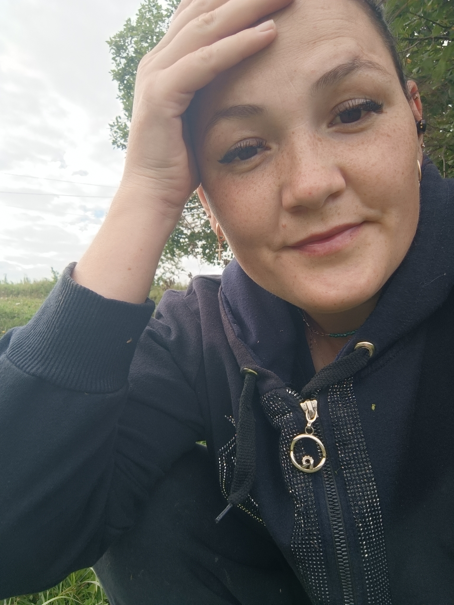 A close-up, low-angle, medium shot captures a woman's face from the chest up, with her right hand on her forehead. She is looking directly at the camera with a slight smile. The woman has fair skin with freckles, dark eyelashes, and dark hair pulled back. She is wearing a dark hooded sweatshirt with a zipper and a beaded necklace. The background consists of trees and a cloudy sky. The location is identified as Karsha, Russia.