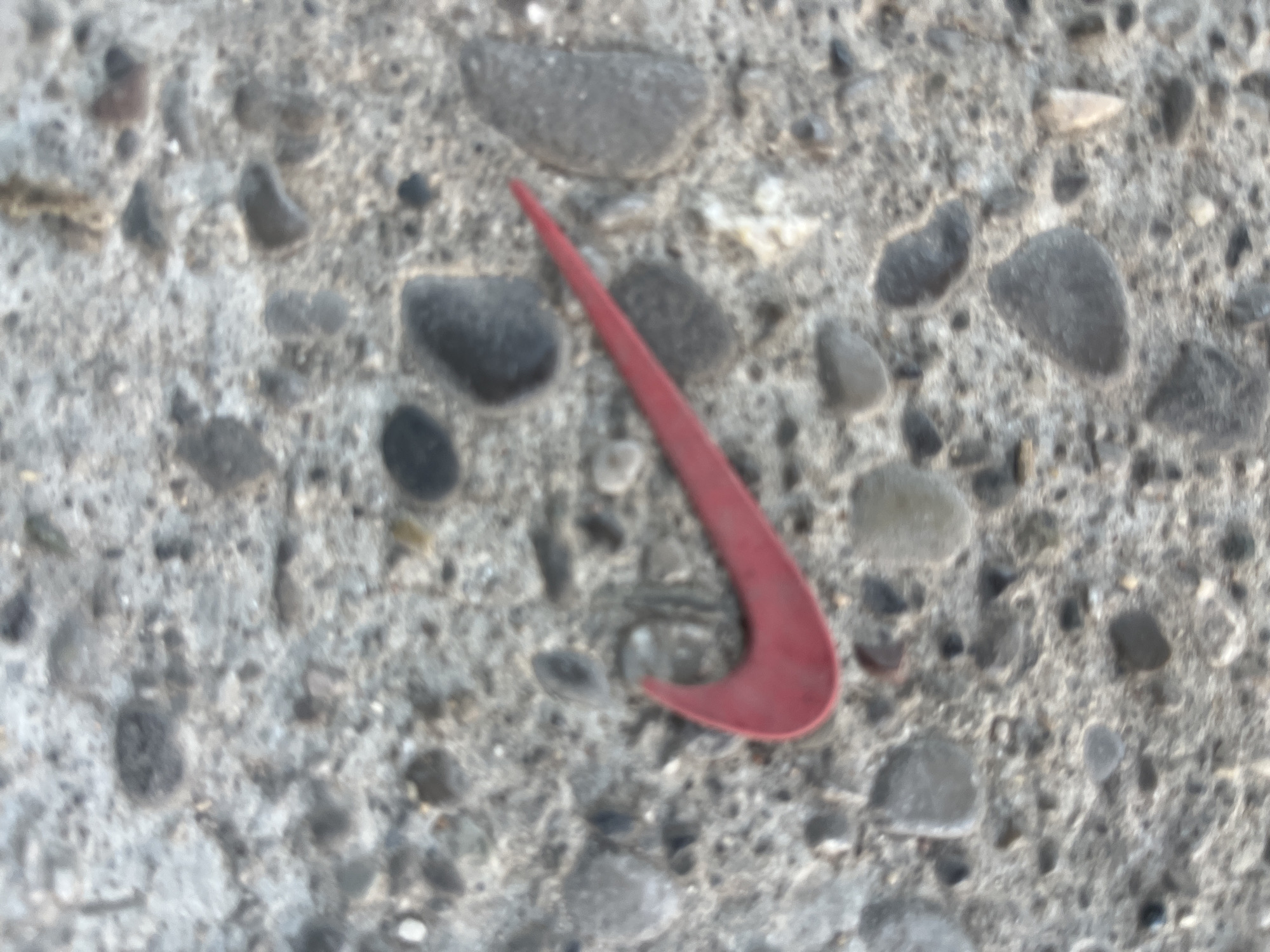 A red Nike swoosh logo is lying on a grey, pebbly asphalt surface. The logo appears to be a detached piece of footwear or accessory. The background consists of irregularly shaped and sized pebbles embedded in the asphalt. There are no people or any other discernible objects or actions in the scene. The image is taken from a top-down perspective.