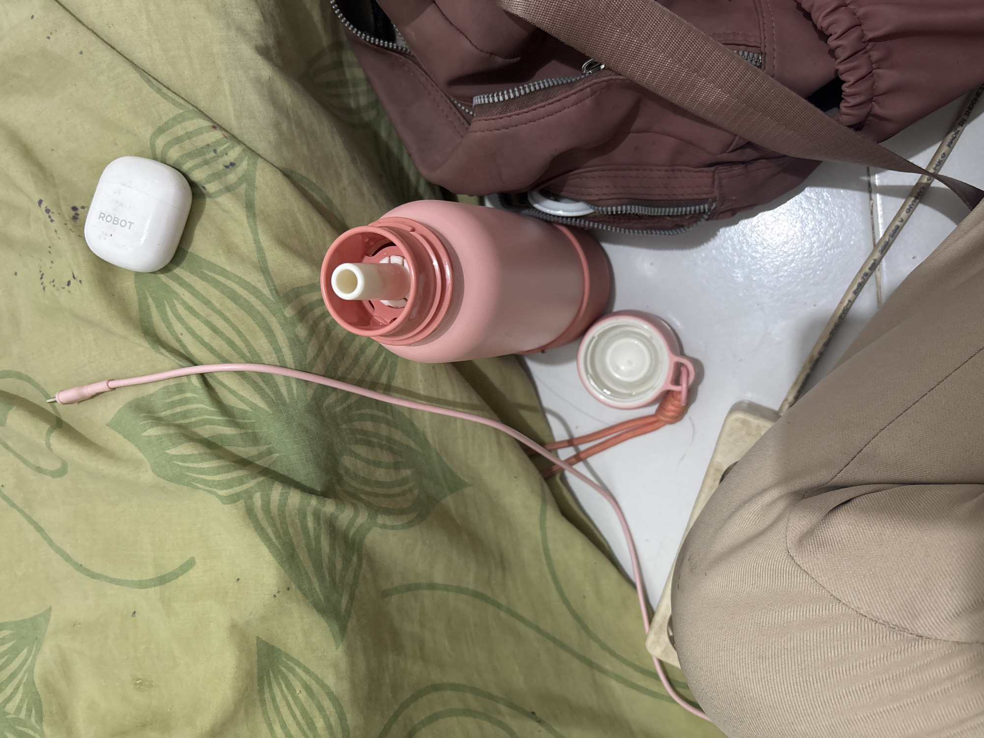 The image depicts a close-up, high-angle shot of personal items arranged on a patterned fabric surface. In the foreground, a person's beige trousers are visible. To the right, a portion of a dark pink bag with zippers and a strap is shown. In the center, a pink water bottle with its cap detached sits next to a white object, likely a wireless earbud case with "ROBOT" printed on it. A pink charging cable extends from the earbud case towards the top left of the frame. The background consists of a light green fabric with a repeating leaf pattern. The floor appears to be tiled. The scene is an indoor environment, likely a bedroom or a living area. There is no indication of the specific location within North Jakarta, Indonesia.