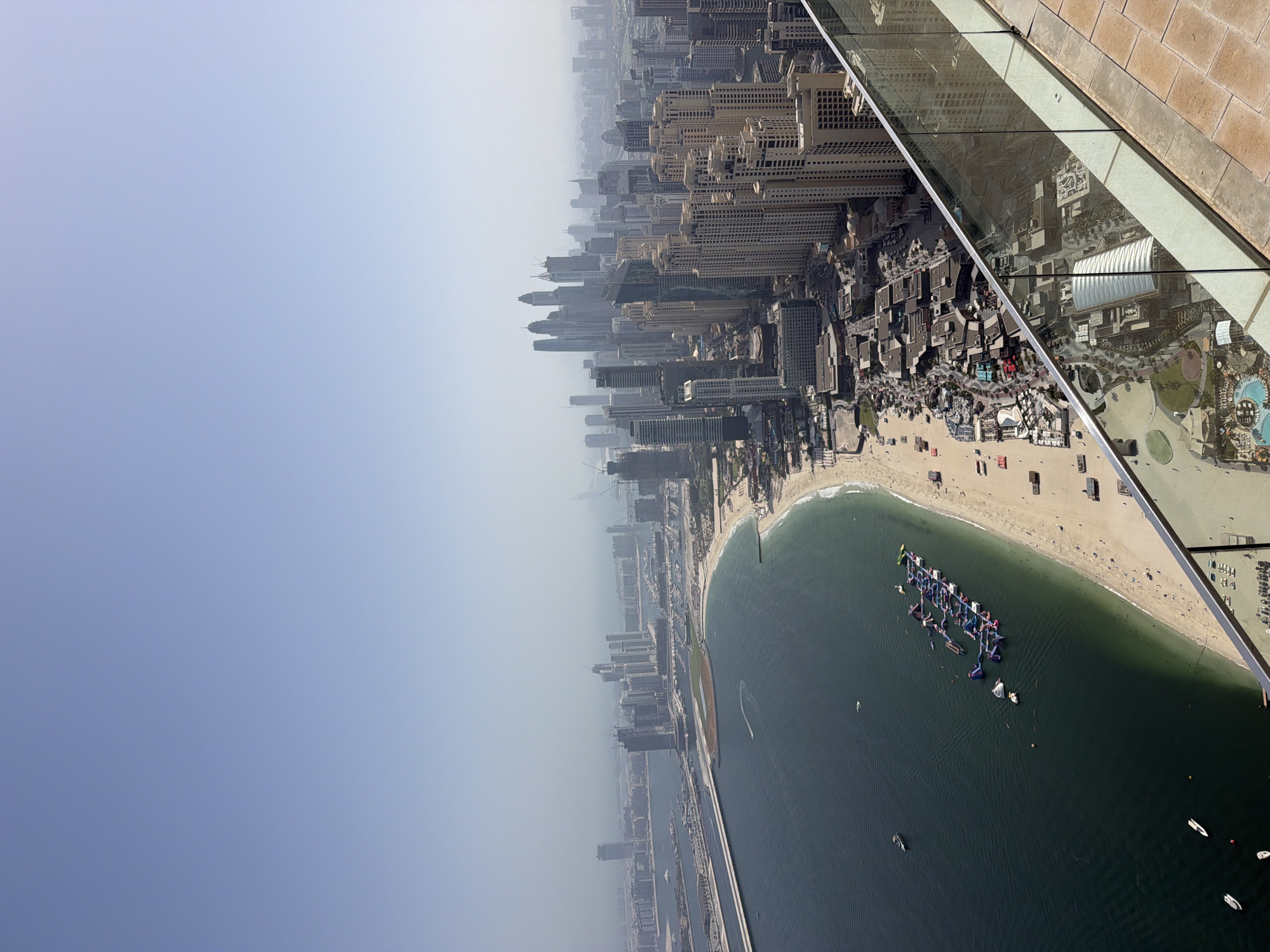 The image depicts an elevated view, likely from a skyscraper observation deck, of Dubai Marina and Jumeirah Beach Residence (JBR) in Dubai, United Arab Emirates. The sky is hazy. Several high-rise buildings are visible along the coastline. The coastline features a sandy beach. In the water, there appears to be a large inflatable structure, likely an aqua park, with some watercraft nearby. People can be seen on the beach. The observation deck's glass panel and railing are visible in the upper right corner of the image, with the city reflected in the glass.