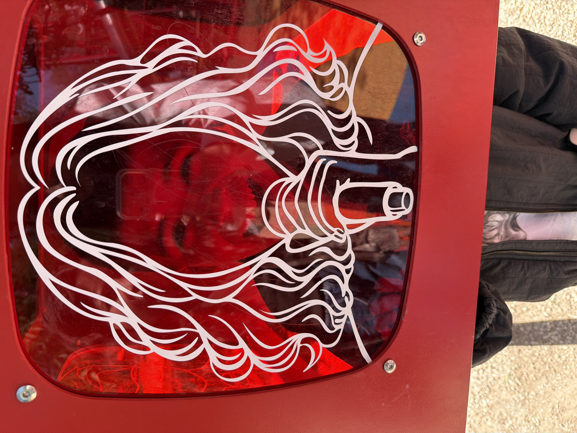 The image shows a stylized, white-line drawing of a lion's head on a red, translucent panel. The panel is rectangular with rounded corners and secured by bolts. The lion's mane is depicted as flowing, wavy lines. Through the translucent panel, indistinct red shapes are visible. A portion of a person's arm, wearing a black jacket and showing a tattoo, is partially visible on the right side of the frame. The background behind the jacket appears to be a light-colored surface, possibly a sidewalk. The location is Yerevan, Armenia.