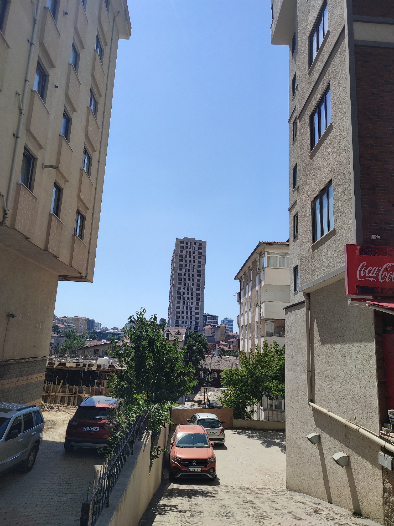 The image depicts a street scene in Kadıköy, Turkey, under a clear blue sky. On the left, a light-colored, multi-story apartment building with numerous windows dominates the foreground. To the right, another apartment building with a textured facade and brick accents is visible, featuring a red Coca-Cola sign. Between these buildings, a narrow street leads down towards a collection of smaller buildings and trees. Several cars are parked on the street, including a silver SUV, a red SUV with license plate "34 PCO 589", and an orange sedan with license plate "34 HAU 676". In the background, a tall, unfinished skyscraper is under construction, and other residential buildings are scattered across the cityscape. The overall impression is of an urban environment with a mix of older and newer construction.