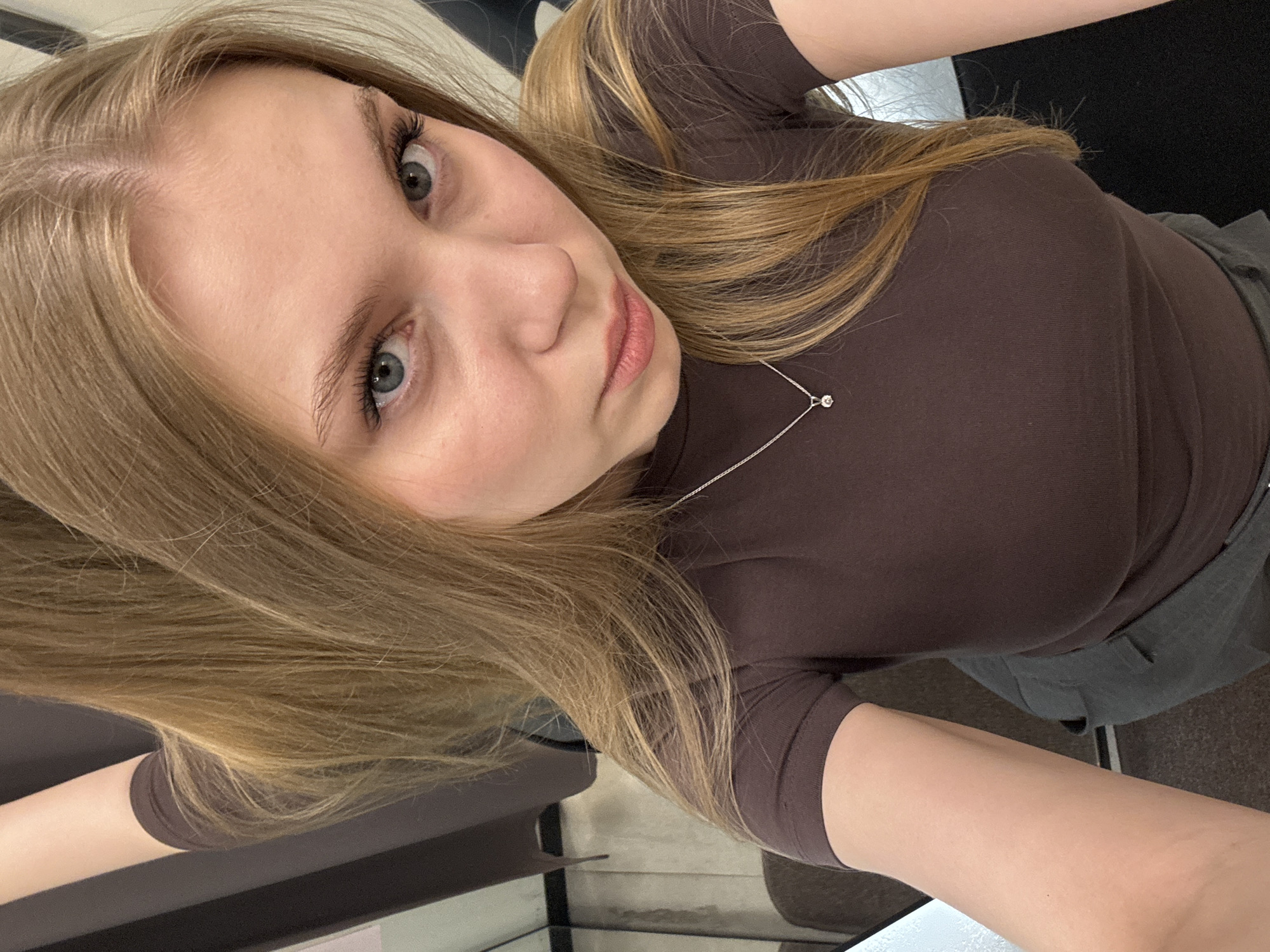 A young, light-skinned woman with long, straight ash blonde hair, parted down the center, looks directly at the camera in a high-angle selfie. Her blue-grey eyes are highlighted with mascara, and her natural-toned lips are slightly parted. She wears a fitted, short-sleeved dark brown top and dark grey trousers. A thin silver chain necklace with a small, single clear stone pendant rests on her chest. The background depicts an indoor, minimalist environment with dark grey vertical panels or curtains and reflections, suggesting a private commercial space such as a changing room.