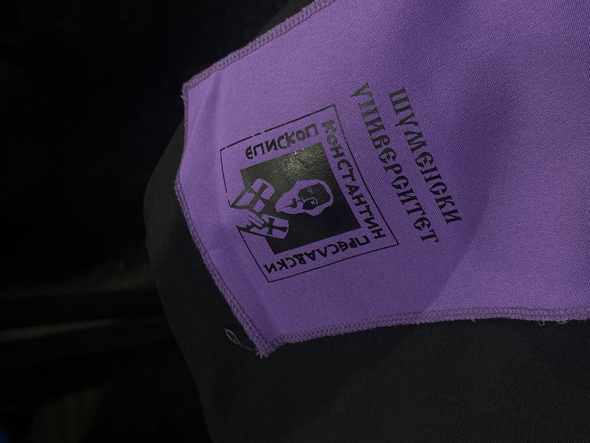 A close-up shot depicts a purple square patch with black text and a logo attached to a black garment or object. The text, in Cyrillic script, spells out "SHUMEN UNIVERSITY 'EPISCOPUS CONSTANTINE PRESAVSKY'." The logo features a stylized black and white representation of a bishop holding religious objects within a square frame. The patch is sewn onto the black fabric. The background is dark and out of focus. The image is likely taken indoors. The location is Shumen, Bulgaria.