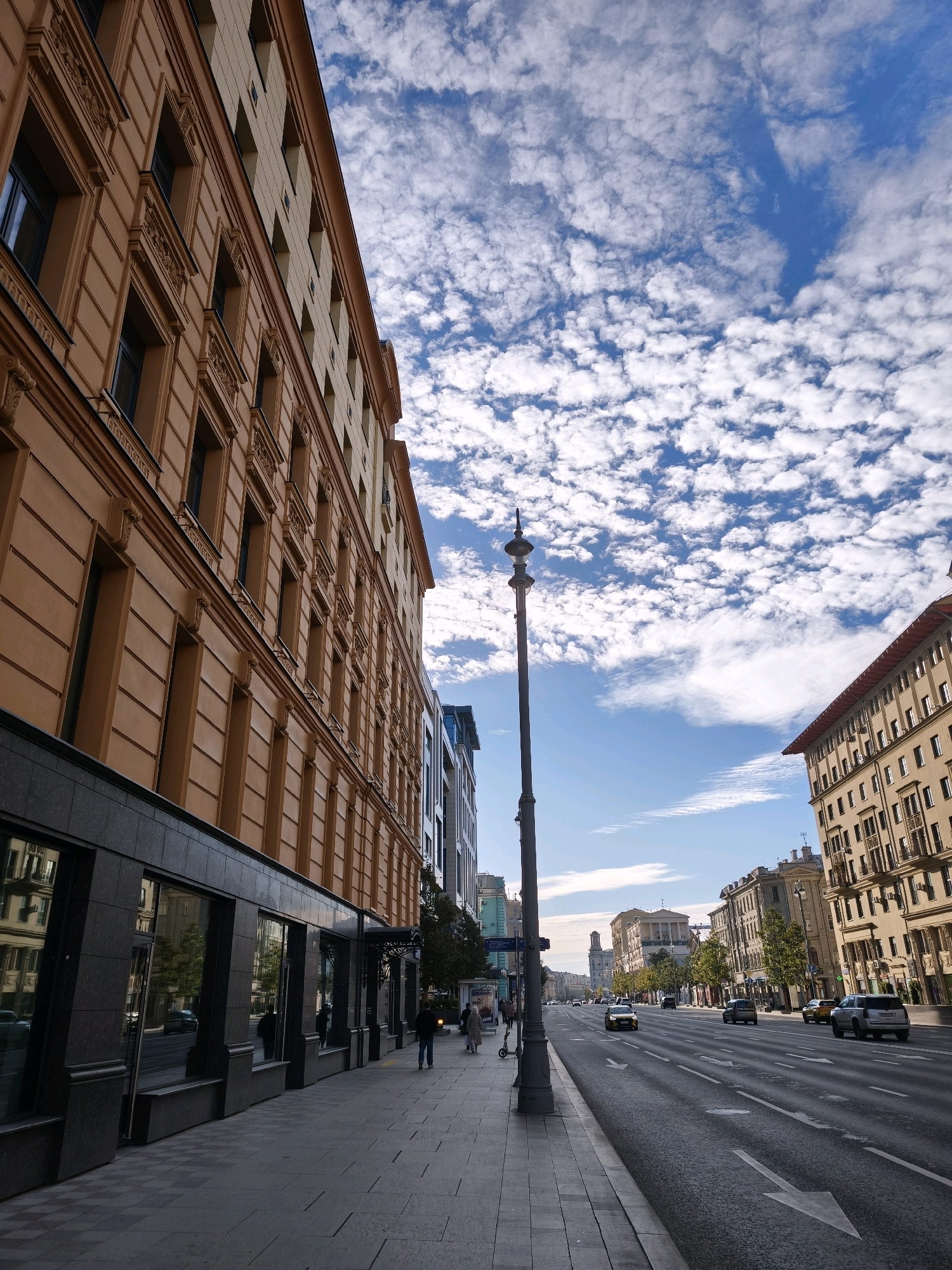 An eye-level, vertical, medium shot captures a wide urban street in Moscow, Russia, under a partially cloudy blue sky. The foreground is dominated by the left side of a tall, beige, classical-style building with ornate architectural details and dark-paned windows. To the right, the street stretches into the distance, lined with more buildings of varying architectural styles, some appearing older and more ornate, others modern. The road has multiple lanes with clear white lane markings and is populated by several cars, including a silver SUV, a yellow taxi, and a dark sedan. A lone street lamp with a decorative finial stands on the sidewalk in the mid-ground. Two individuals are visible walking on the sidewalk in the middle distance, one in dark clothing and another in lighter clothing. The reflection of buildings and greenery is visible in the dark, polished facade of the building in the immediate foreground, suggesting a commercial ground floor. The overall atmosphere is one of a bustling city street during daylight.