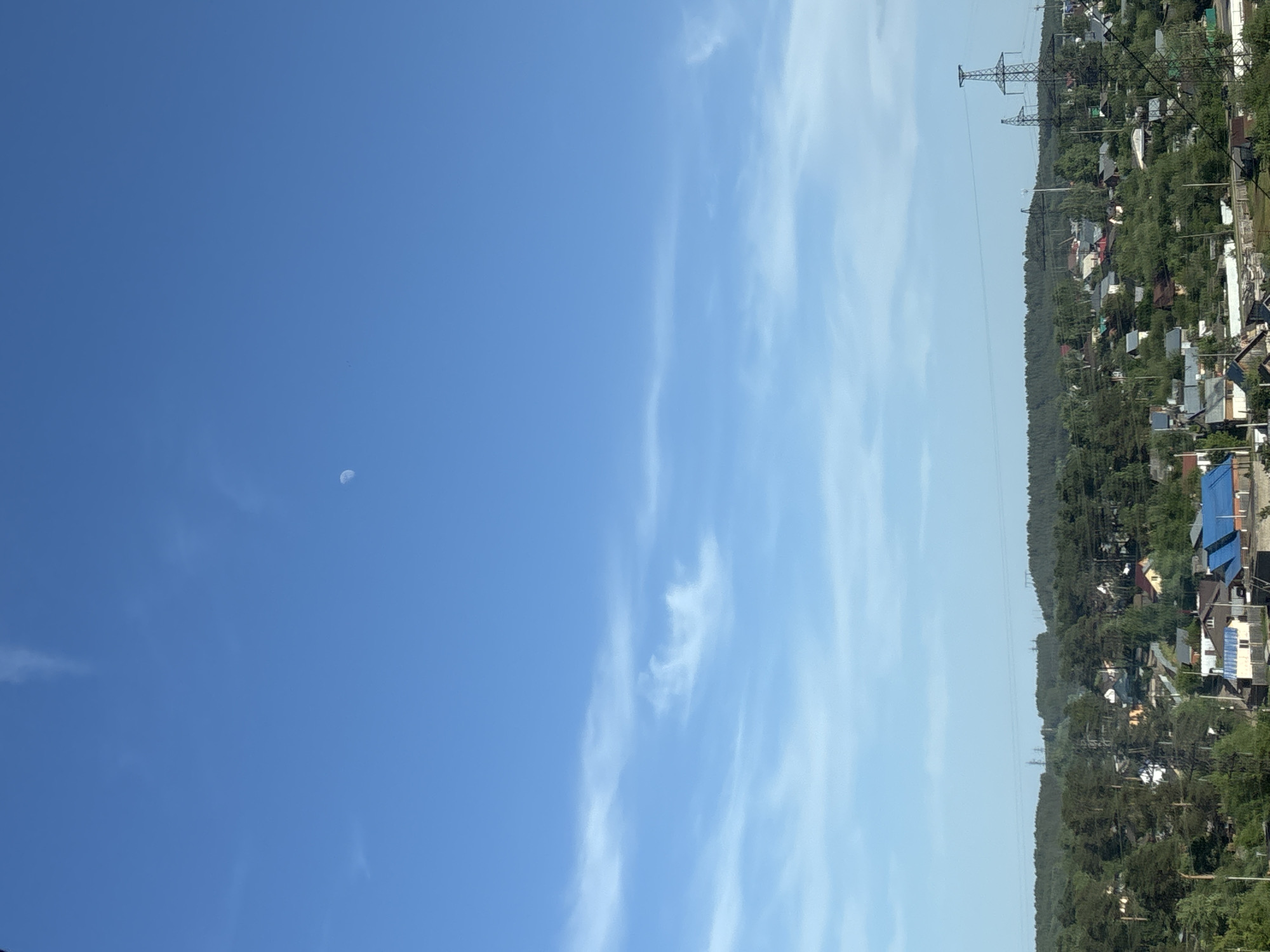 The image shows a daytime scene. The sky is divided into two shades of blue, with a brighter, cloudless blue on the left side and a paler blue with streaks of white clouds on the right. A small crescent moon is visible in the darker blue sky. On the right edge of the image is a landscape of trees and houses, possibly a residential area. Power lines and pylons are visible extending above the trees. The location is in the city of Unknown, Unknown.
