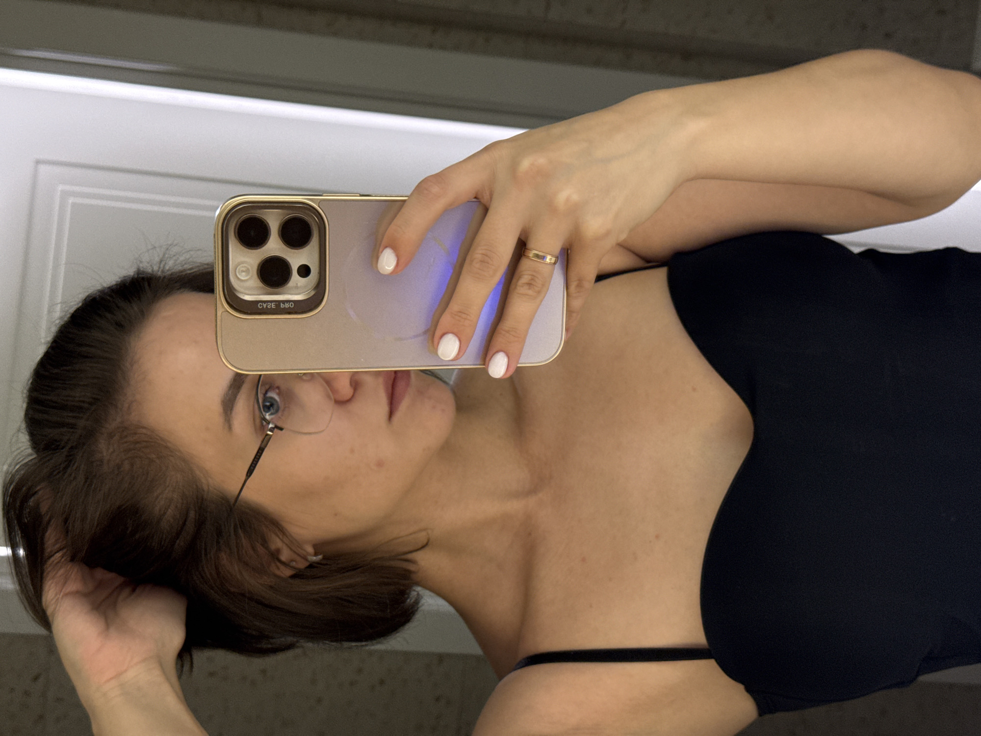 A woman with short brown hair, glasses, and white nail polish is taking a selfie in a bathroom with her phone. She is wearing a black top. The location in Pervomaysky, Russia is undetermined.