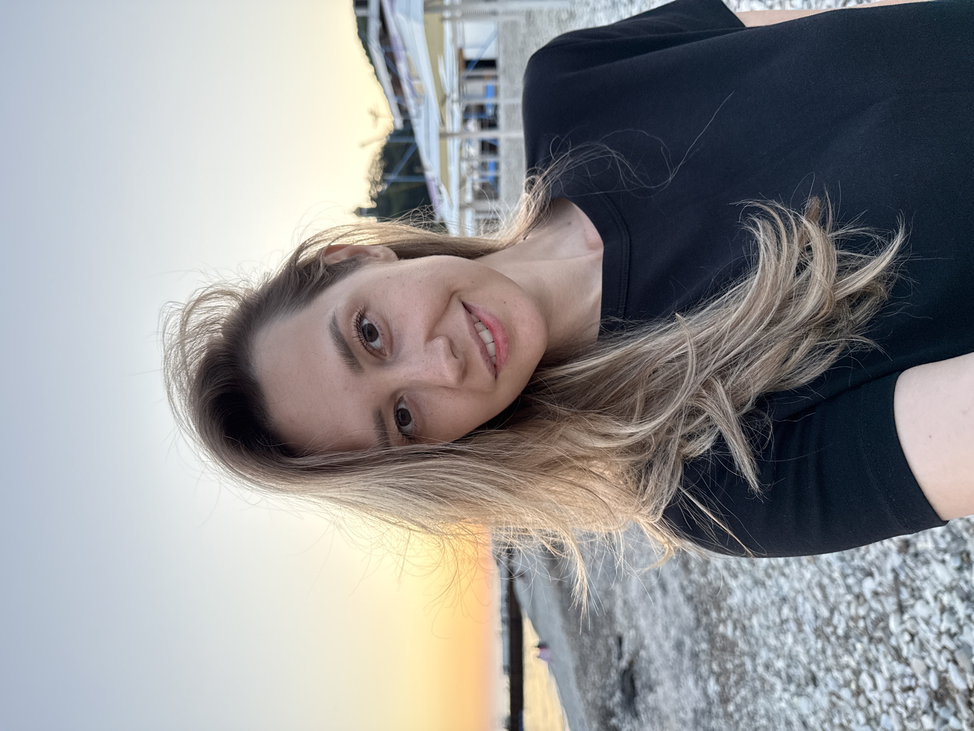 A medium shot shows a woman with blonde hair and fair skin wearing a black t-shirt, standing on a pebble beach at sunset. Her hair is windswept, and she is smiling at the camera. The background features the sea, a beach with scattered people, and some beach umbrellas and buildings. The overall atmosphere is relaxed and casual. The scene is set in Nebug, Russia, indicated by the architecture and the style of beach infrastructure.