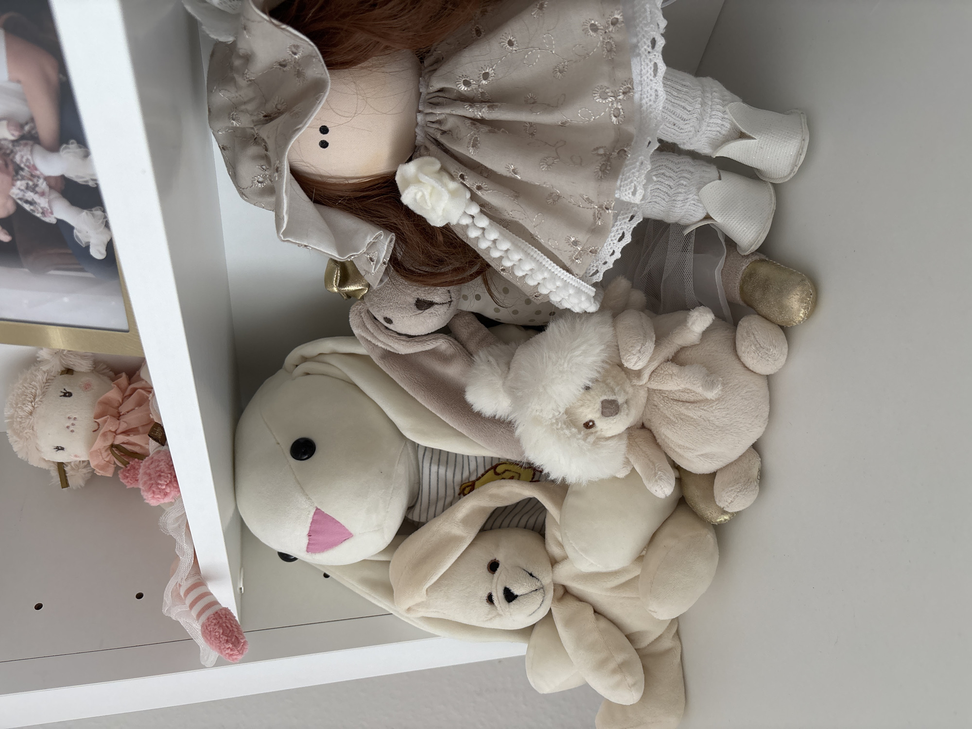 The scene depicts an interior view of a white shelving unit in the city of Unknown, Unknown. The upper shelf holds a small, light-pink plush toy with a ruffled collar and striped pink and white legs positioned on the left. Adjacent to it on the right is a gold-framed photograph, partially visible, showing the lower body of an infant or small child, wearing a floral dress and white ruffled leggings, being held by an unseen individual. The lower shelf is occupied by several plush toys. A large white rabbit with black button eyes, a pink triangular nose, and long floppy ears is centrally located, wearing a grey and white striped shirt with a yellow emblem. To its left, a smaller cream-colored plush rabbit with floppy ears and black stitched features leans against the larger rabbit. A brownish-grey plush rabbit with a gold bow near its ear is partially visible behind the cream-colored rabbit. In the front-center, a small, fluffy, light-cream plush animal with brown eyes and a light-brown nose rests. To the right, a fabric doll with reddish-brown hair, black stitched eyes, and a light-colored face stands. The doll is dressed in a beige gown with embroidered floral patterns and white lace trim, adorned with a white pom-pom string terminating in a white fabric rose on its chest. The doll wears white ribbed socks and white shoes with pointed toes. Partially visible beneath the doll's attire are a pair of golden shoes associated with another plush toy.