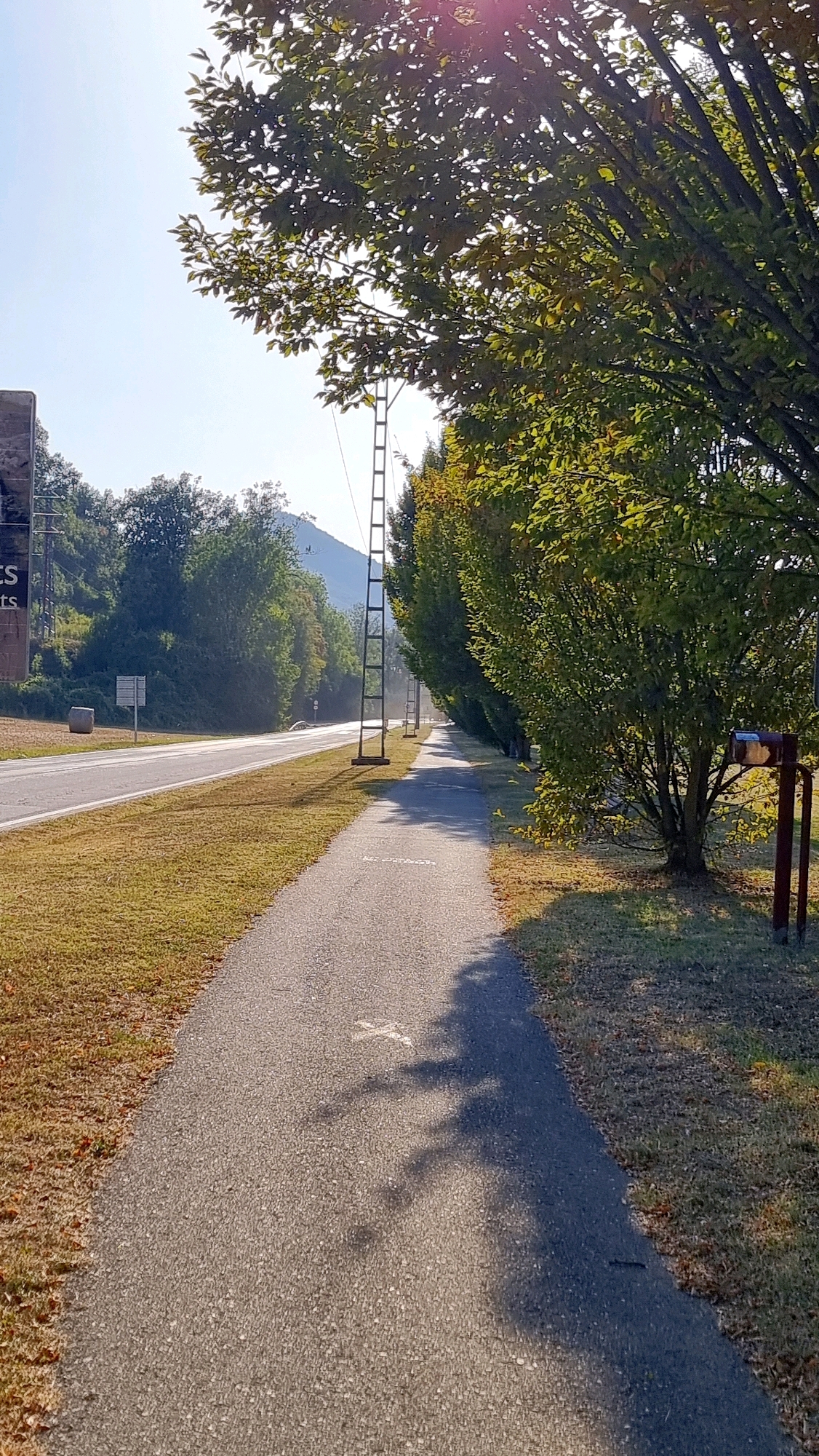 The image depicts a paved pathway running alongside a road, with trees lining the right side of the pathway. The pathway appears to be a pedestrian or bike path. In the background, there are more trees and a hill with a structure on it. The sky is clear and blue. There are some shadows on the pathway, suggesting it is a sunny day. The scene appears to be in a rural or suburban area. A roadside sign is visible on the left side of the road. There is also a hay bale in the field next to the road. A mailbox is present on the right side of the pathway. The overall impression is one of a peaceful, natural setting.