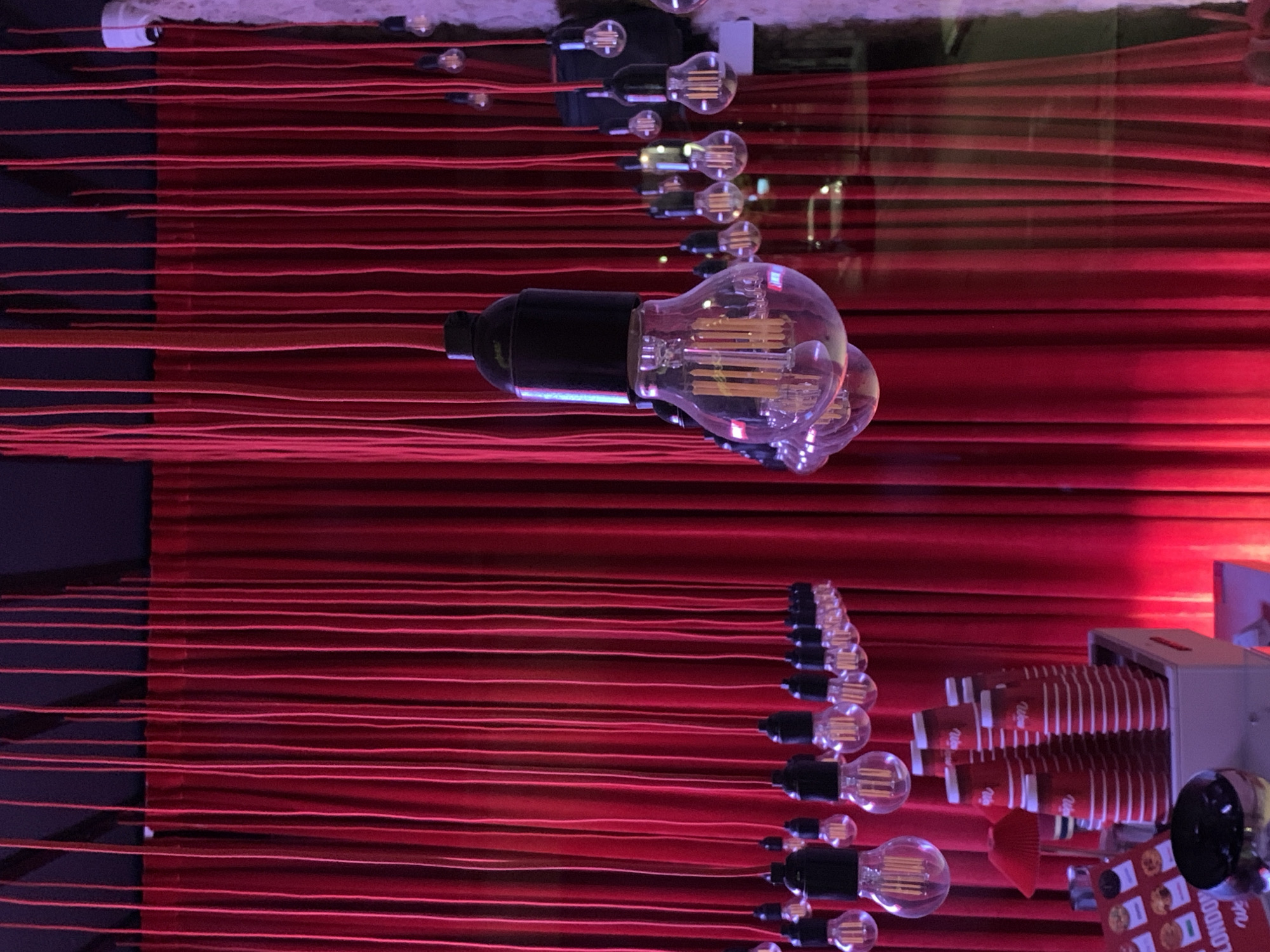 A tilted, horizontally oriented shot features a unique display. Red strings stretch across the frame, interspersed with light bulbs and dark-colored bulb bases. Additional objects include a box of red cups. The scene is in Tel-Aviv, Israel.