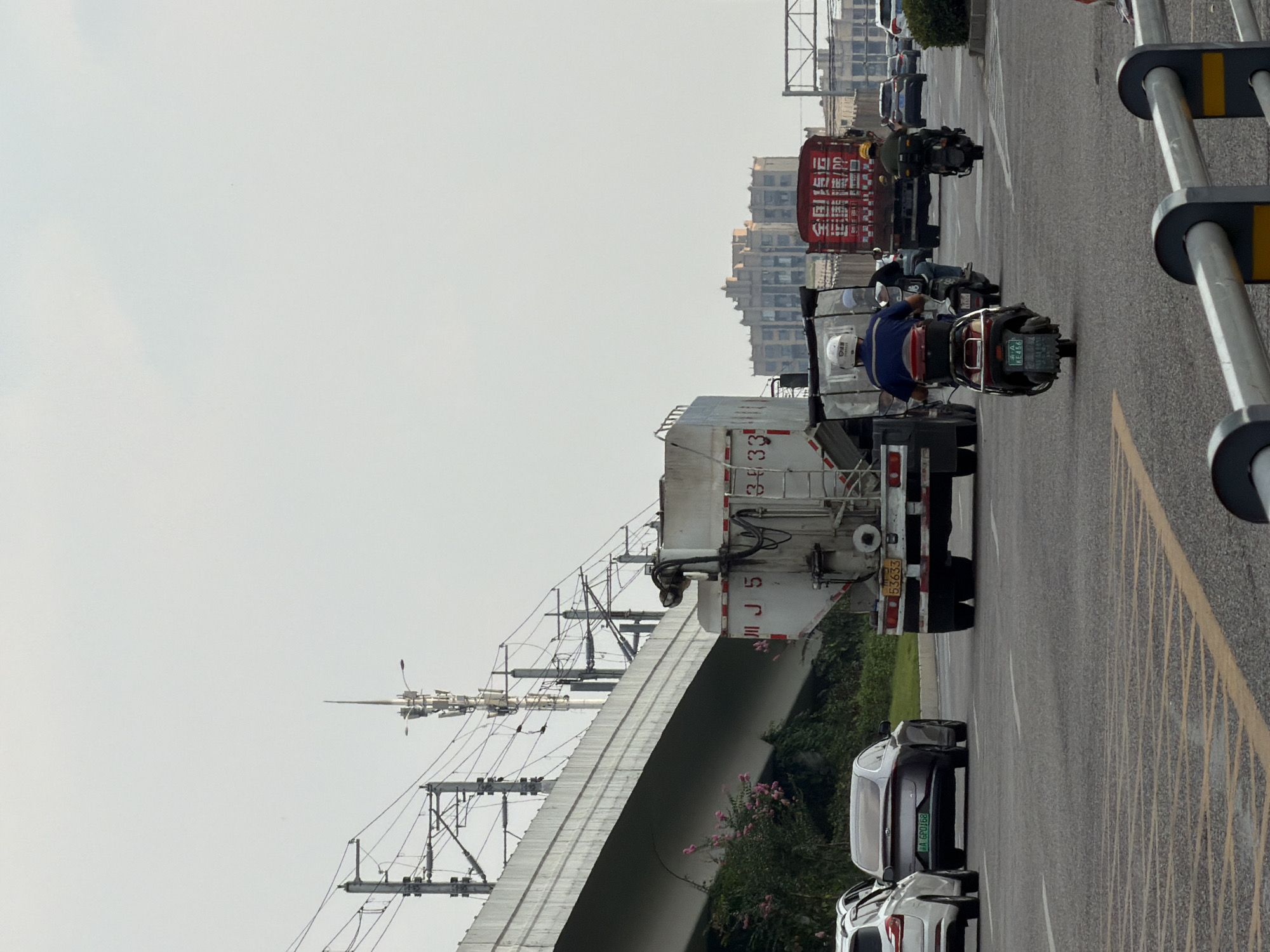 In Jiangjin, China, on a clear day, traffic flows along a multi-lane road beneath an elevated highway structure. The road surface is asphalt, marked with white lane lines and yellow diagonal warning lines. On the highway above, electrical infrastructure for what appears to be a train line is visible, with multiple poles and catenary wires. To the left, pink flowers bloom on a vegetated embankment beneath the elevated structure.
The road is populated with various vehicles. In the foreground, a motorcyclist in a blue shirt and white helmet rides a scooter. Behind him, another scooter carries a passenger, also wearing a helmet. Further ahead, a large white truck with a cylindrical tank and red and white markings is visible, traveling in the same direction. To the left of this truck, a dark-colored sedan with a license plate reading "A GP0168" is driving. Another white vehicle is partially visible to the far left.
In the distance, a red truck with white Chinese characters on its side is also moving along the road. Several other vehicles, including cars and what appear to be more trucks, are visible in the background, along with a building complex with a distinctive tiered architectural style. The sky is overcast with a pale grey hue.