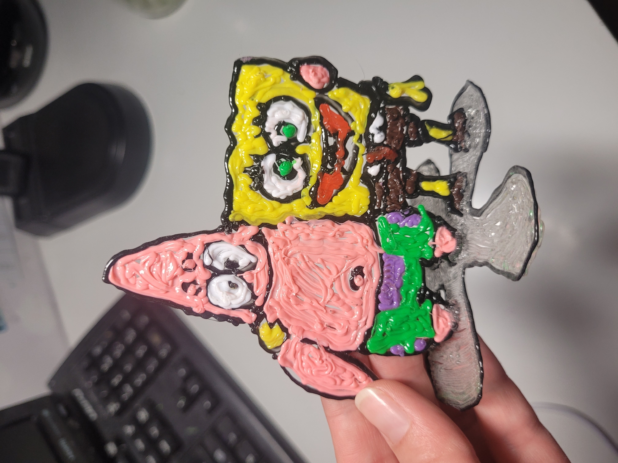 A hand holds up a colorful 3D pen creation depicting SpongeBob SquarePants riding Patrick Star. SpongeBob, made from yellow plastic, is positioned atop Patrick. He has prominent white eyes with bright green pupils, a red, open mouth, and is wearing brown shorts with a yellow top. Patrick, rendered in pink plastic, forms the base, with white eyes and black pupils, and is dressed in green shorts with purple accents. The figures are outlined with black plastic. A translucent, glittery grey structure, possibly resembling a surfboard or a wave, is attached beneath Patrick.
The creation is held over what appears to be a light-colored desk or workspace. In the blurred background to the bottom left, a dark-colored keyboard is partially visible. To the top left, a black, out-of-focus desk lamp or similar object is present. The overall texture of the figures clearly shows the extruded lines typical of 3D pen art. The scene takes place indoors, likely at a desk in Izhevsk, Russia. No visible text is present on the object itself.