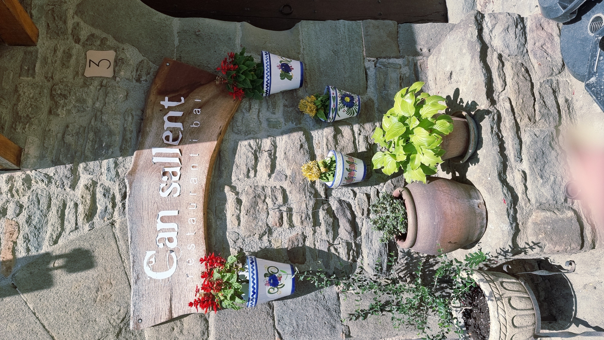 The image shows the exterior facade of a stone building in Rupit, Spain. A weathered wooden sign with white lettering reads "Can sallent restaurant i bar". Several potted plants are displayed on the stone wall and on stone ledges. These include red flowers in a pot with blue and white trim, yellow flowers in two small decorated pots, a plant with bright green leaves in a small pot, and a larger terracotta pot containing a green plant. To the right of the sign, a square beige plaque with the number "3" is affixed to the stone wall. A dark doorway is visible on the far right of the image. The scene is illuminated by sunlight, casting shadows on the stone wall.