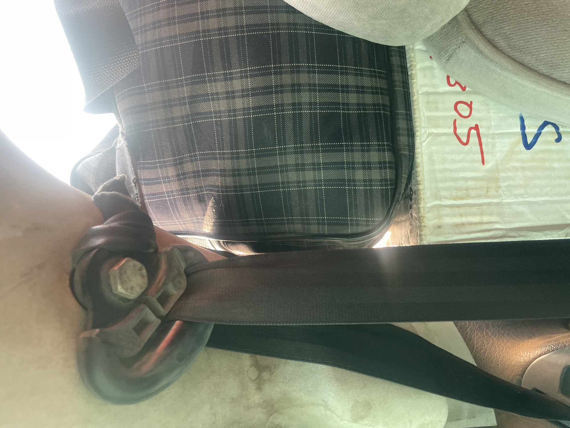 The image shows a close-up of a black seatbelt fastened to a metal anchor point. The anchor point is secured by a large bolt. To the right of the seatbelt is a portion of a dark grey and white plaid bag. Below the bag, there is a white cardboard box with red and blue writing on it. The red writing appears to be "SOS", and the blue writing is an incomplete "S". The scene appears to be inside a vehicle, likely an older model, given the texture of the surrounding surfaces which suggest worn upholstery or interior panels. The overall lighting suggests a bright, possibly sunny day, with a strong glare in the upper right portion of the image. The location is identified as Wauro Jabbe, Nigeria.