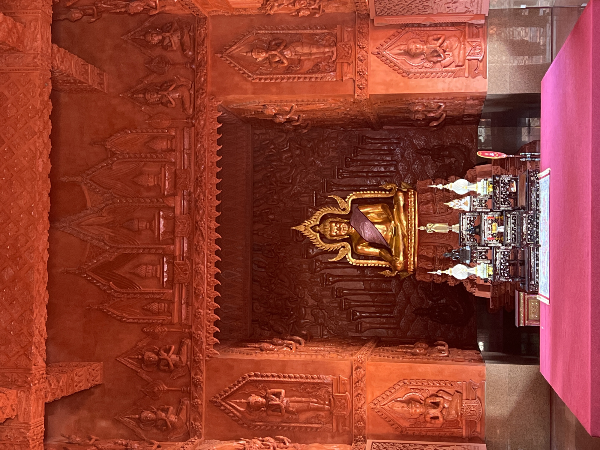 This is a full-frame, eye-level, medium shot of the interior of a temple. In the center of the image, a golden statue of the Buddha is seated in front of a dark, ornate wall carving. The Buddha statue wears a purple cloth draped over its shoulder. In front of the Buddha statue, there is a decorative altar with smaller statues and religious items. The entire interior of the temple is a vibrant orange-red color, with intricate carvings and architectural details covering the walls and ceiling. These carvings depict figures and patterns, adding to the rich and detailed atmosphere of the space. In the foreground, a large, red-covered platform occupies the lower portion of the frame. The overall scene is one of religious reverence and ornate artistry within a temple setting.