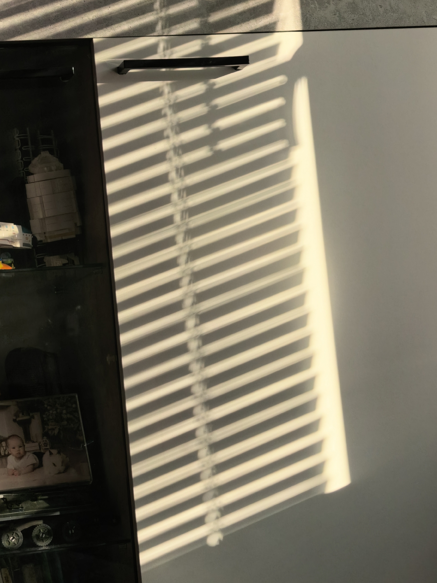 The image shows a section of a dark glass-fronted display cabinet next to a white wall, both partially covered by the shadows of window blinds. Inside the cabinet, various objects are visible on multiple shelves. The lowest level shows items stacked on top of what appear to be vintage toys and baby pictures. A partially visible object looks like a Lego building set on an upper shelf. The right third of the image is dominated by the shadow pattern cast by window blinds onto the plain white wall. The room appears to be inside a home. There are no humans in the frame. The image does not provide information about the city of Sterlitamak, Russia.
