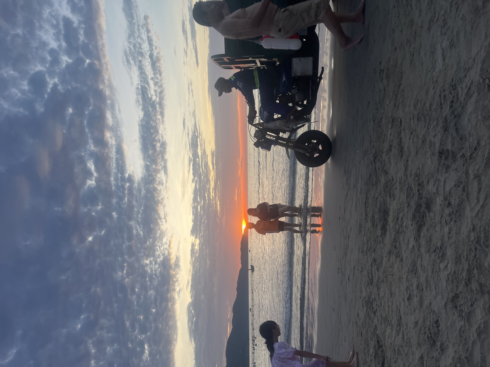 This image captures a sunset scene on a beach in Đà Nẵng, Vietnam. The sky is partially cloudy, displaying a gradient from deep blue at the top to vibrant orange and yellow hues at the horizon where the sun is setting over the water. Distant mountains are visible on the left side of the horizon. The sea is calm with small waves gently reaching the sandy shore.
In the foreground, a young girl in a light-colored dress is visible on the far left, bending over with her back partially to the viewer, near the water's edge. Further right, two individuals, likely women, are standing partially submerged in the shallow water, silhouetted against the bright sunset reflection; one wears an orange top, the other a darker one, and their reflections are visible on the wet sand.
A man in a uniform, consisting of a cap and a blue long-sleeved shirt with reflective stripes, is seated on and operating a motor-tricycle (a modified motorcycle with a cargo bed) on the wet sand. He is facing right, moving along the water's edge. On the far right, partially cropped, another man is walking barefoot on the sand, shirtless and wearing light-colored shorts. Several small boats are scattered across the distant water, visible as dark silhouettes against the bright horizon. The sandy beach is damp and reflects the sky's colors in the foreground.