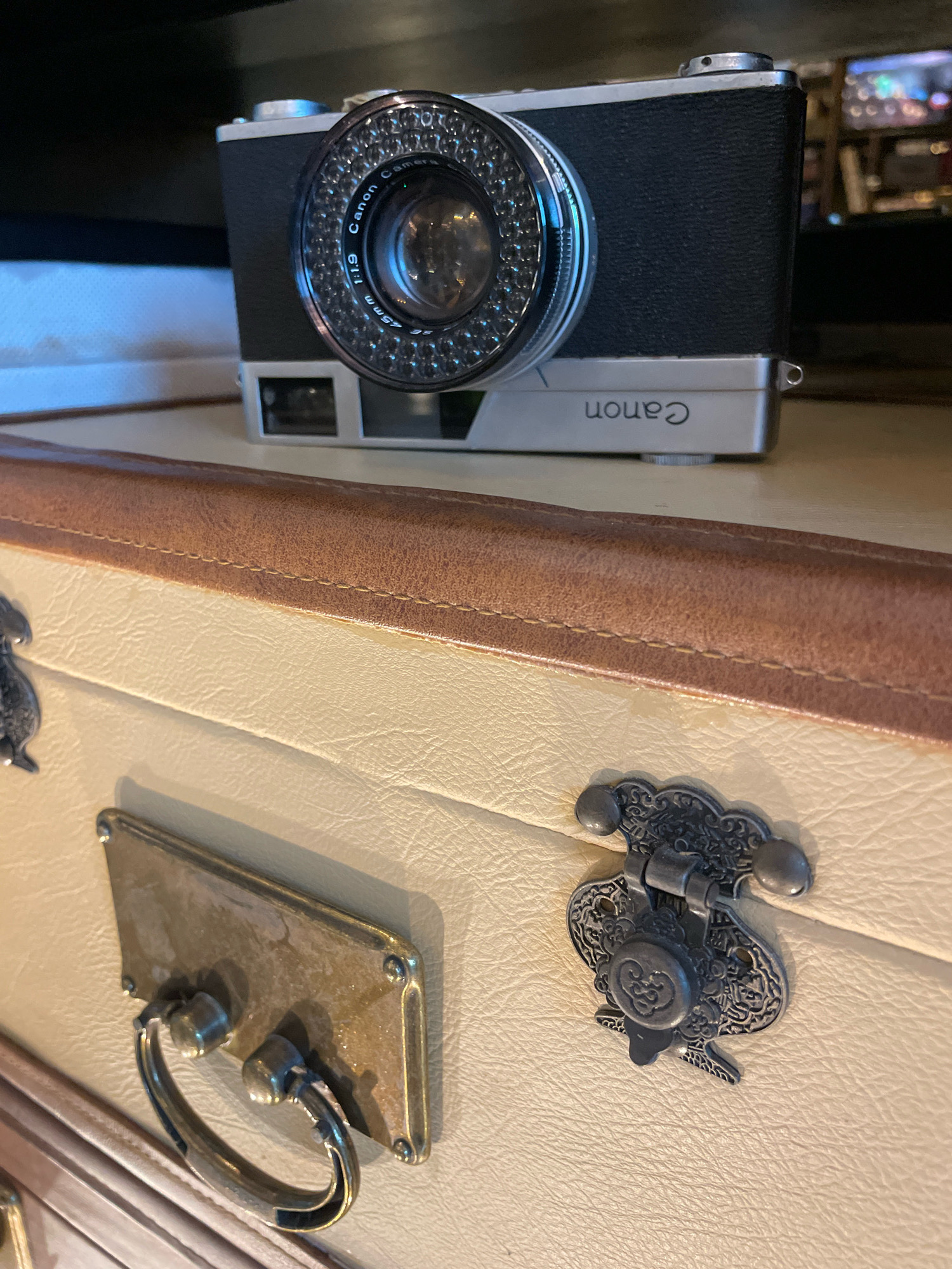 A vintage Canon camera rests on top of a cream-colored suitcase with brown leather trim. The suitcase has ornate silver hardware, including a decorative latch and a rectangular plate with a handle. The camera, with a black textured body and a silver lens ring, is positioned on the top surface of the suitcase. The background is dark and out of focus, with a hint of blurred lights in the distance, possibly indicating an indoor setting like a shop or bar. The scene appears to be set in Pattaya City, Thailand, though no specific landmarks or contextual clues are visible within the image itself to confirm this location.
