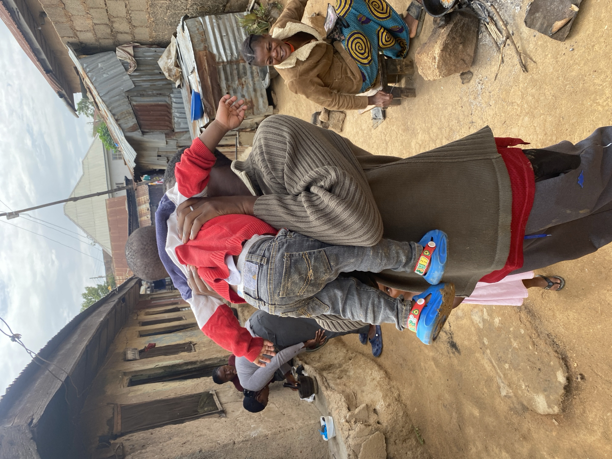 The image shows an outdoor scene in Jos, Nigeria. A person is holding a child wearing a red and white sweater and gray jeans. The child is wearing blue shoes with a red and green pattern. Behind them, a woman in a brown jacket and patterned skirt is cooking over an open fire. Other people are visible in the background near a building. The ground is dirt. There are corrugated metal structures nearby.