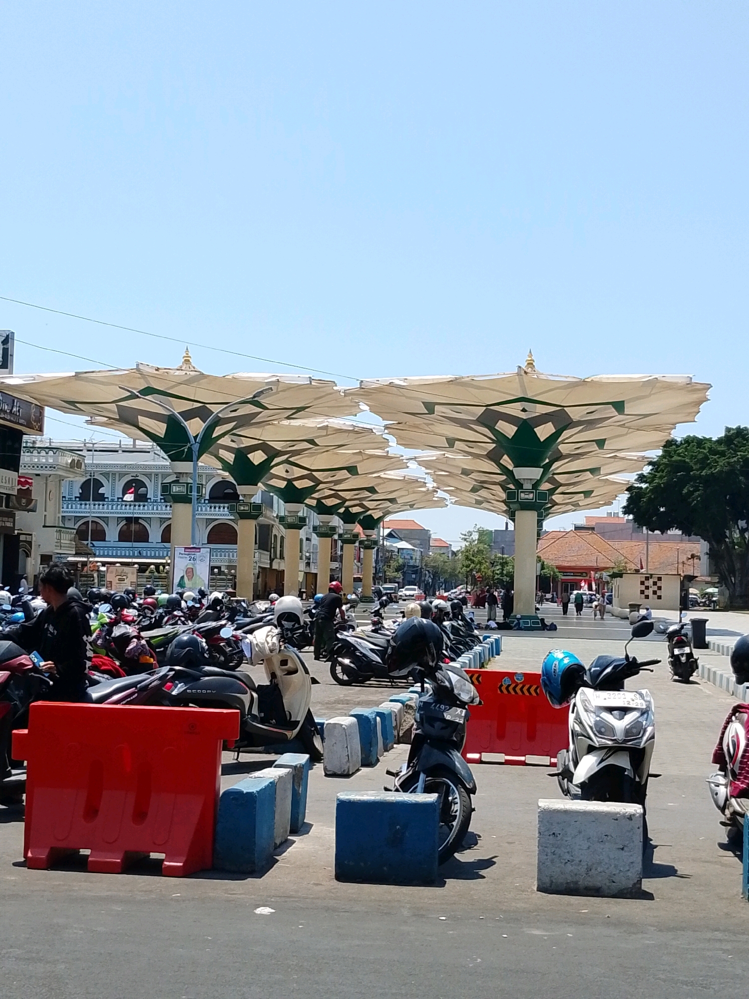 The image depicts an outdoor parking area filled with numerous parked motorcycles under a canopy of large, decorative, umbrella-like structures. These structures are supported by pillars and are a prominent feature of the scene. In the background, there are buildings with classical architectural elements, including arched windows and columns. Several people are visible, some standing and others walking, amidst the parked vehicles and in the distance. The ground is paved, and there are some red and blue barriers present. The sky is clear and blue, indicating a sunny day. This scene is identifiable as the area around the Great Mosque of Pasuruan (Masjid Agung Pasuruan) in Pasuruan City, East Java, Indonesia, due to the distinctive umbrella-like canopies which are a well-known feature of this location, providing shade for congregants and visitors.