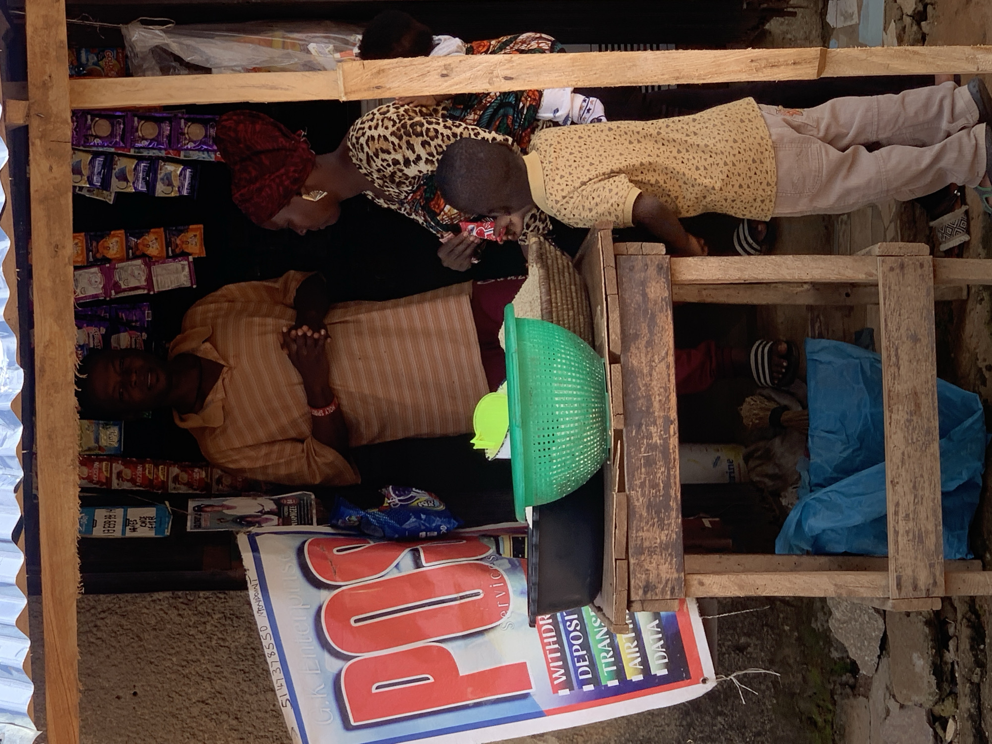 In Sabon Tasha, Nigeria, a small open-air shop displays a variety of goods on shelves. A man in a striped shirt stands behind the counter, his hands clasped. To his right, a woman with a red headscarf and a leopard-print top interacts with a young boy. The boy, wearing a patterned yellow shirt and tan pants, stands facing a wooden table laden with items, including a green colander filled with rice and a dark bowl. A large sign for "G.K Enterprise POS Services" is propped up on the left side of the scene, listing services such as withdrawals, deposits, and data. The shop is constructed with wood and corrugated metal roofing, and the ground is a mixture of dirt and stones. Bags of goods and miscellaneous items are visible within the shop's interior.