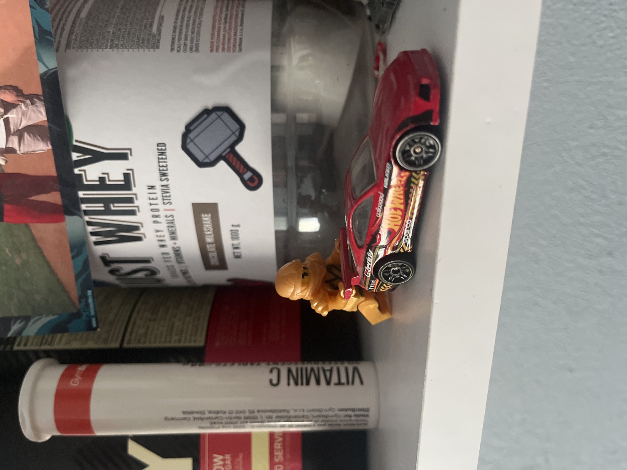 The image shows a shelf with various items, including a white bottle with red and blue accents, a large white container with "JUST WHEY" written on it, and a red toy car with a figure of a character pushing it. The white bottle has text that appears to be in German and English, including "Gym" and "VITAMIN C". The large container is a protein supplement, and its label features an illustration of a hammer. The toy car is a Hot Wheels brand, with racing decals.
The provided context mentions the city of Pokrovnik, Bulgaria, but there are no discernible elements within the image that specifically place it in that location. The scene appears to be an indoor setting, likely a shelf in a home or store, given the arrangement of products. The lighting is natural and appears to be coming from the left. The composition is a close-up shot, with the shelf and its contents filling the frame. There are no people visibly present in the image, only the toy figure. The focus is on the objects and their arrangement, suggesting a still-life or product photography context.
