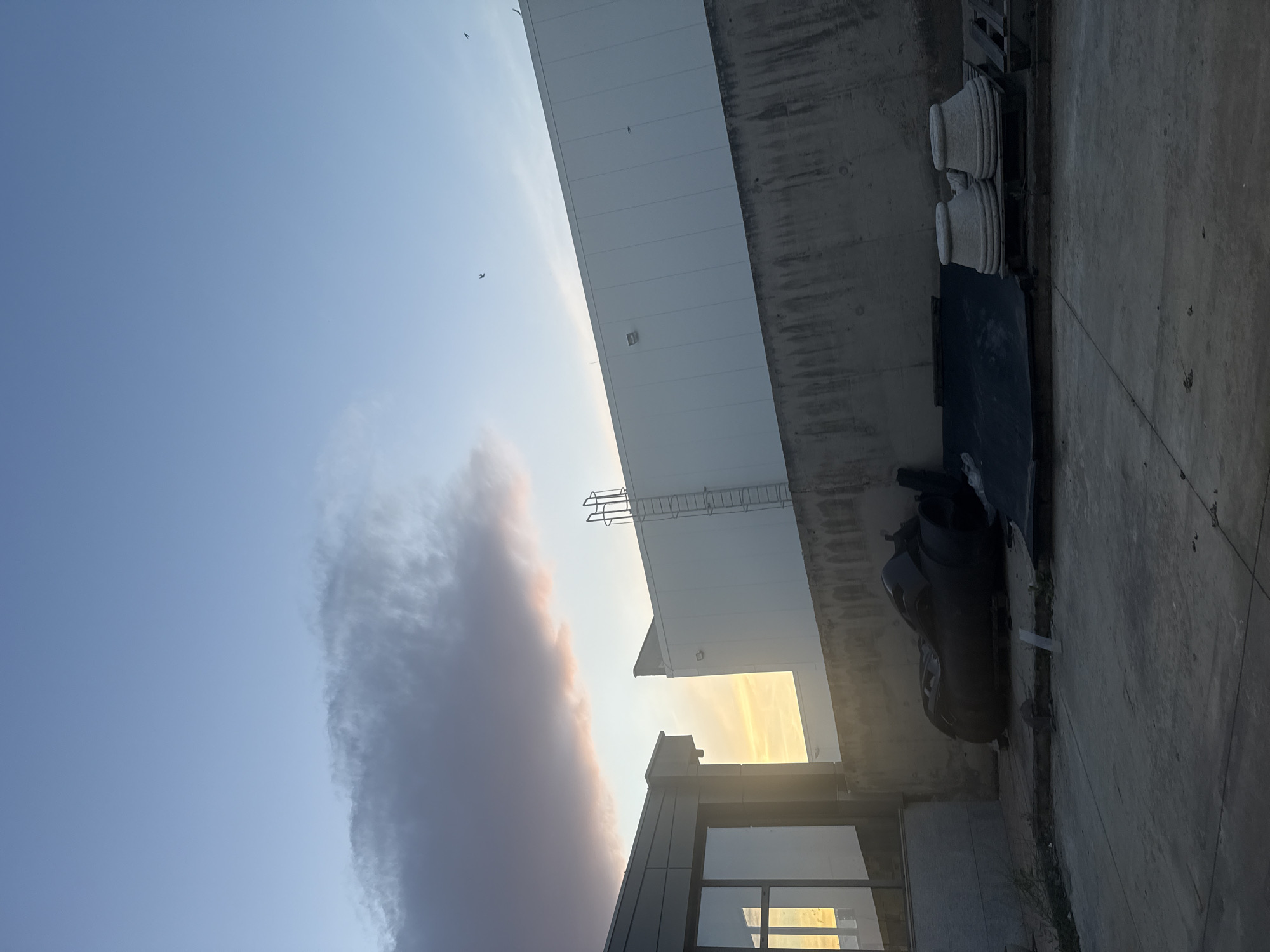 The image shows an outdoor urban scene in Pishtigovo, Bulgaria. The sky is blue with a large cloud. Part of a large, white, industrial building is visible. A metal ladder is attached to the side of the building. A gray concrete wall runs alongside the building. There are decorative planters and other objects. A modern building with glass windows is visible at the bottom of the frame. The lighting suggests it is either sunrise or sunset.