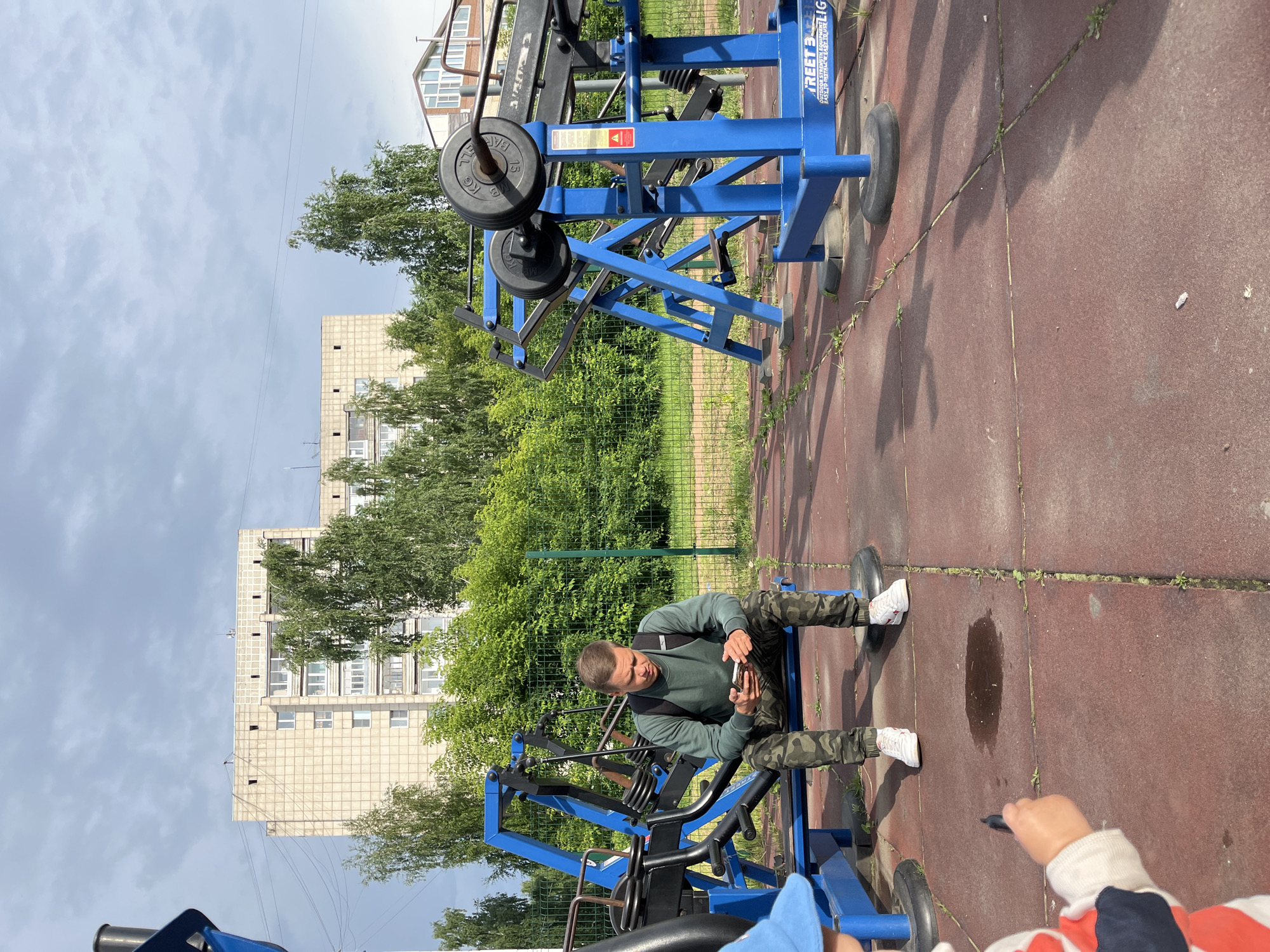 In Chaikovsky, Russia, on a cloudy day, a young man sits on a blue weight training apparatus, engrossed in his mobile phone. He wears a green long-sleeved shirt and camouflage pants, with a backpack resting on his shoulders. White athletic shoes adorn his feet. The outdoor gym equipment, painted a vibrant blue, features metal weights and bars. The ground is paved with red rubber tiles, interspersed with green grass. In the background, a multi-story concrete residential building stands tall, surrounded by lush green trees and a wire fence. Further to the right, another building with a pitched roof is visible. A child, partially visible in the foreground, holds a dark object, possibly a toy, and wears a blue hat and a striped shirt.