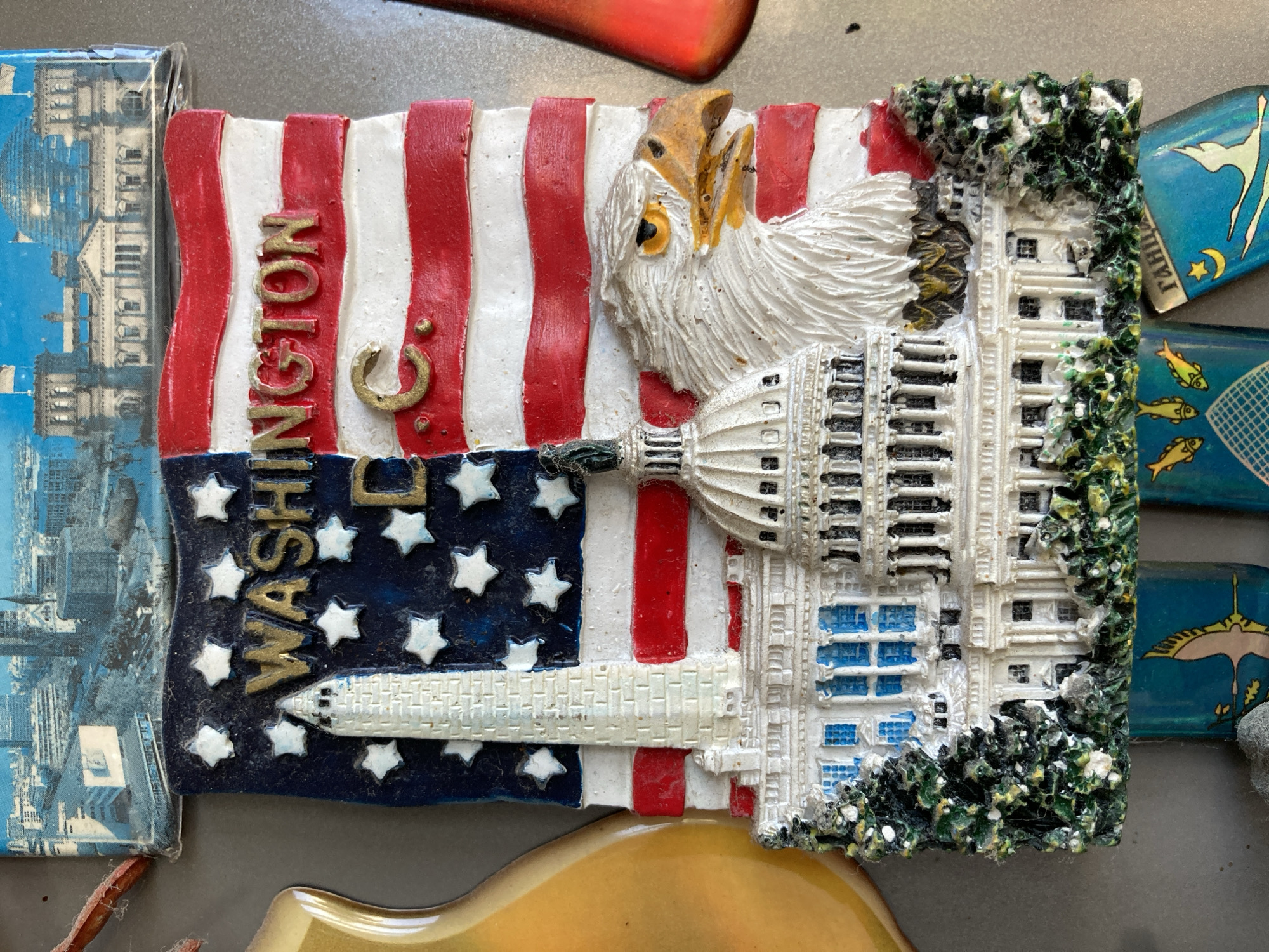 The image is a close-up of a souvenir magnet designed to represent Washington D.C. The magnet features a depiction of the American flag with the words "WASHINGTON D.C." in gold lettering. Superimposed on the flag are iconic Washington D.C. landmarks: the Washington Monument and the United States Capitol Building. An eagle's head is prominently placed on the right side of the magnet, facing right. The bottom portion of the magnet shows stylized greenery and what appear to be small, teal-colored tabs with images of fish and a crane, as well as Cyrillic text. The magnet is positioned on a flat, metallic surface, likely a refrigerator. The overall impression is of a collectible item showcasing national symbols and landmarks.