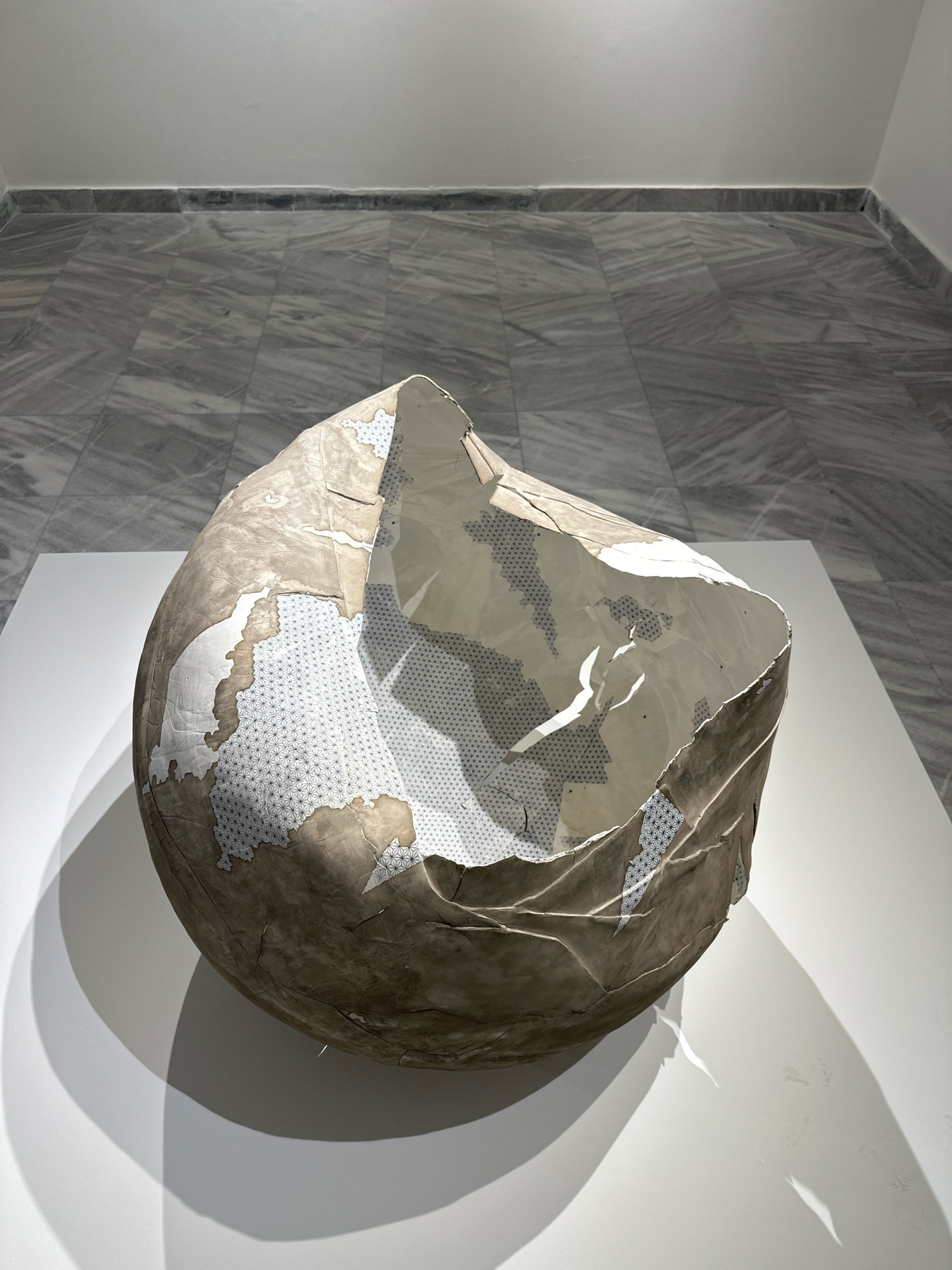 A large, abstract sculpture resembling a crumpled paper orb is displayed on a white pedestal in an art gallery. The sculpture's exterior is a rough, textured brown paper, with sections peeling away to reveal an interior lined with white paper featuring a repeating pattern of blue geometric shapes. The sculpture casts a prominent shadow on the white pedestal and the adjacent wall. The background consists of a wall and a floor with a gray and white marbled tile pattern. The setting appears to be a modern art exhibition space. There are no people visible in the image. This artwork is located in the Belgrade Art Gallery (which is the colloquial name for the National Museum in Belgrade, Serbia).