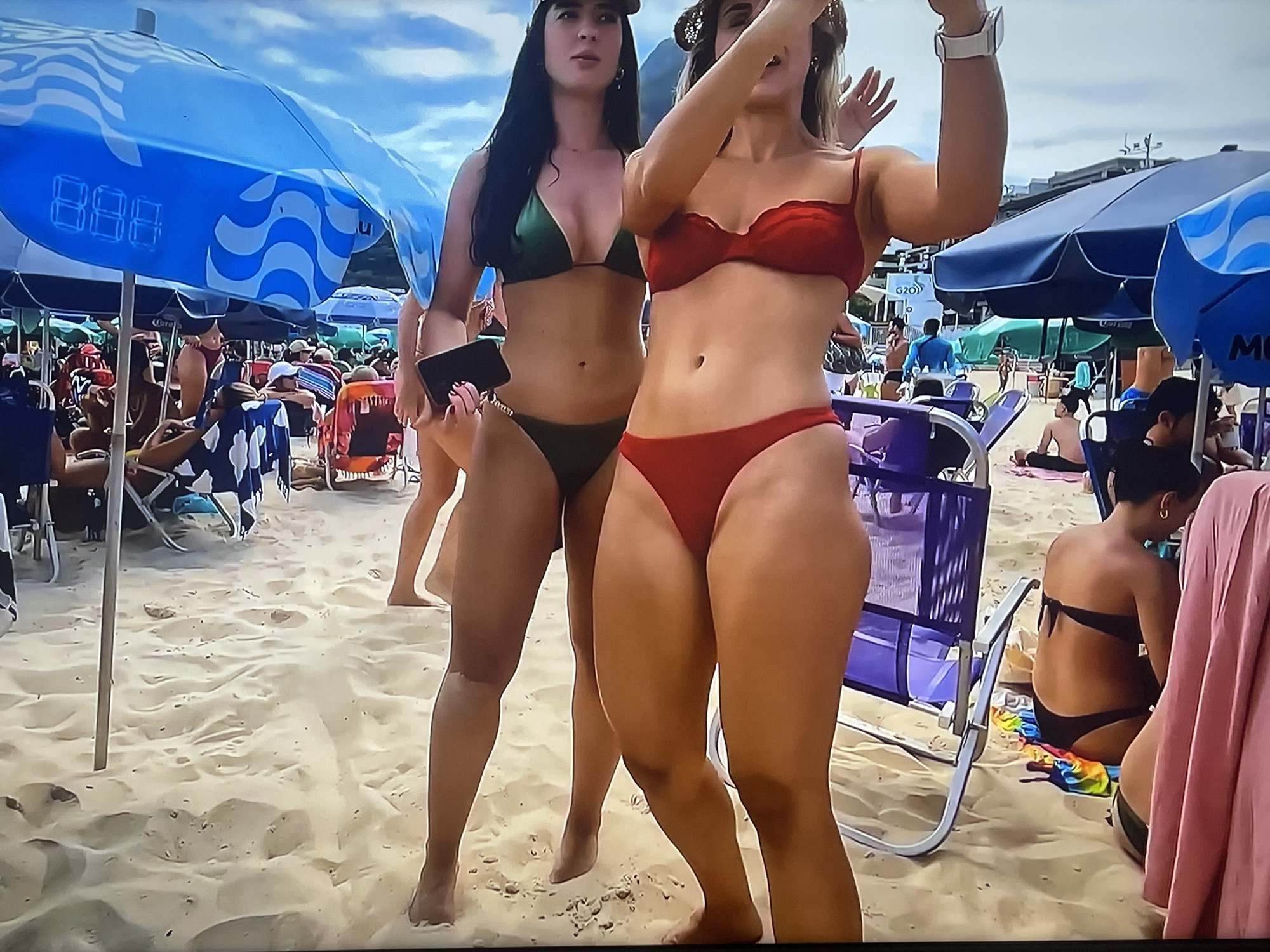 The image depicts a beach scene. In the foreground, two women are visible, both wearing bikinis. The woman on the left is wearing a green bikini and holding a black phone. The woman on the right is wearing a red bikini and appears to be taking a selfie. The beach is sandy, with other beachgoers and umbrellas visible in the background. The sky is cloudy. The location is identified as the city of Shakhovskaya, Russia.