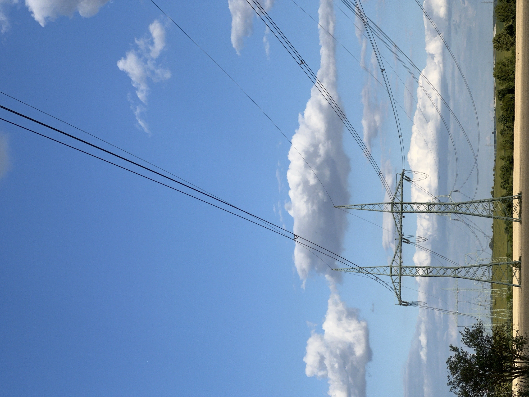 This media file shows a landscape scene with power lines stretching across a bright blue sky dotted with fluffy white and grey clouds. The power lines are supported by tall, green lattice pylons, two of which are prominently featured in the foreground and mid-ground. The pylons are connected by thick black cables that crisscross the sky.
In the background, a green, rolling landscape is visible with a line of trees and some distant structures that appear to be more power pylons. The sun is shining, suggesting it is daytime. The weather appears fair with scattered clouds, creating a dynamic sky. There are no people visible in the media file. The scene evokes a sense of vastness and industrial infrastructure within a natural setting. The location is specified as Újezd u Brna, Czechia. There is no visible text in the media file.