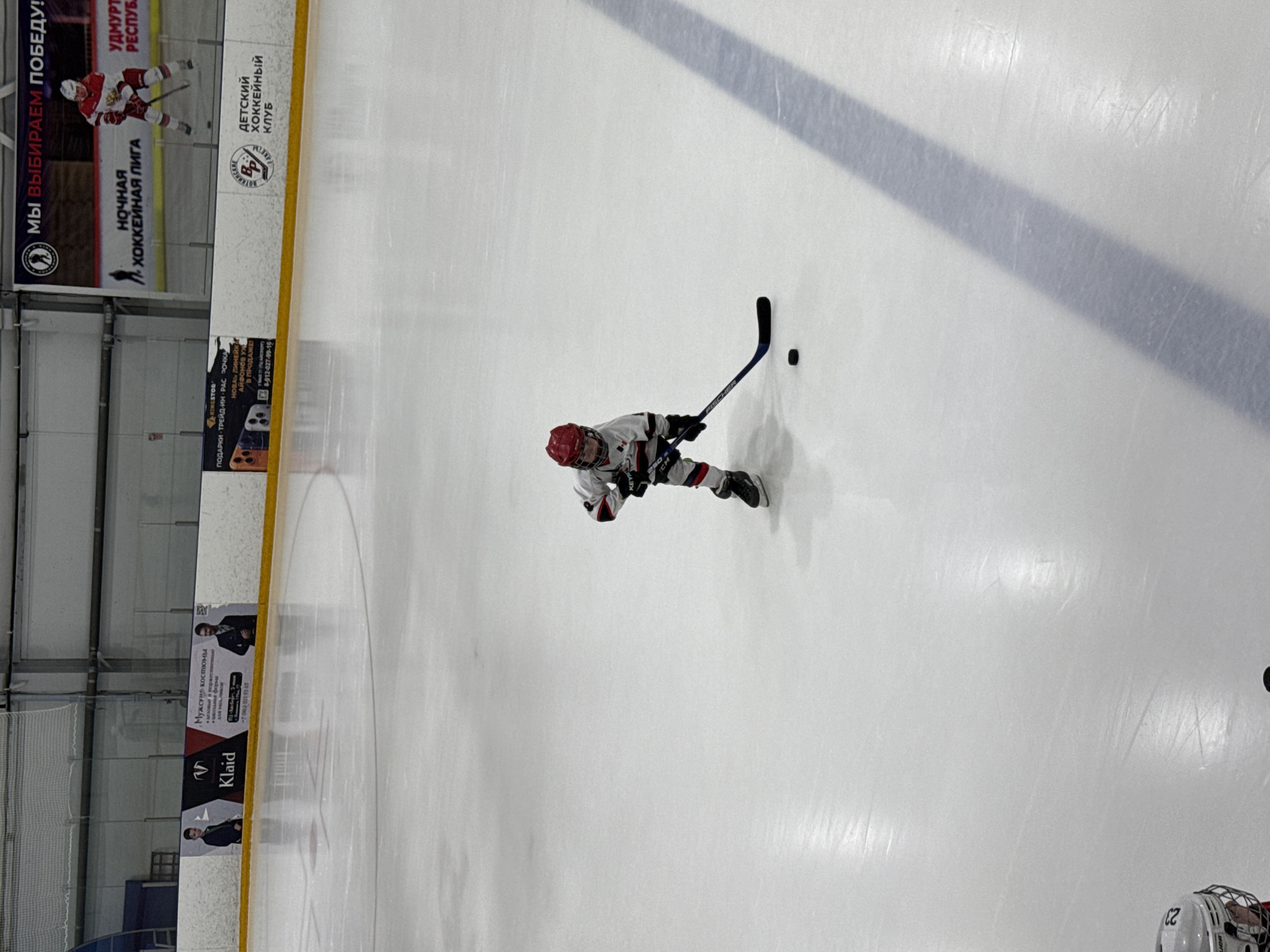 This media file depicts a young boy playing ice hockey on an indoor rink. He is dressed in a white and red hockey uniform, complete with a helmet and gloves, and is in the process of controlling the puck with his stick. The scene is illuminated by overhead lights, suggesting it is daytime or the rink is well-lit.
The background shows advertisements along the rink's boards. One prominent ad in Russian mentions "Votkinsk," "Udmurt Republic," and "Children's Hockey Club," indicating the location. Other advertisements display "Night Hockey League" and various retail businesses, including "Klaid" and "King Stor," which seems to offer gifts and possibly phone sales. The overall atmosphere appears to be focused on the sport and local community engagement. The ice surface shows clear skate marks, evidence of previous play.
