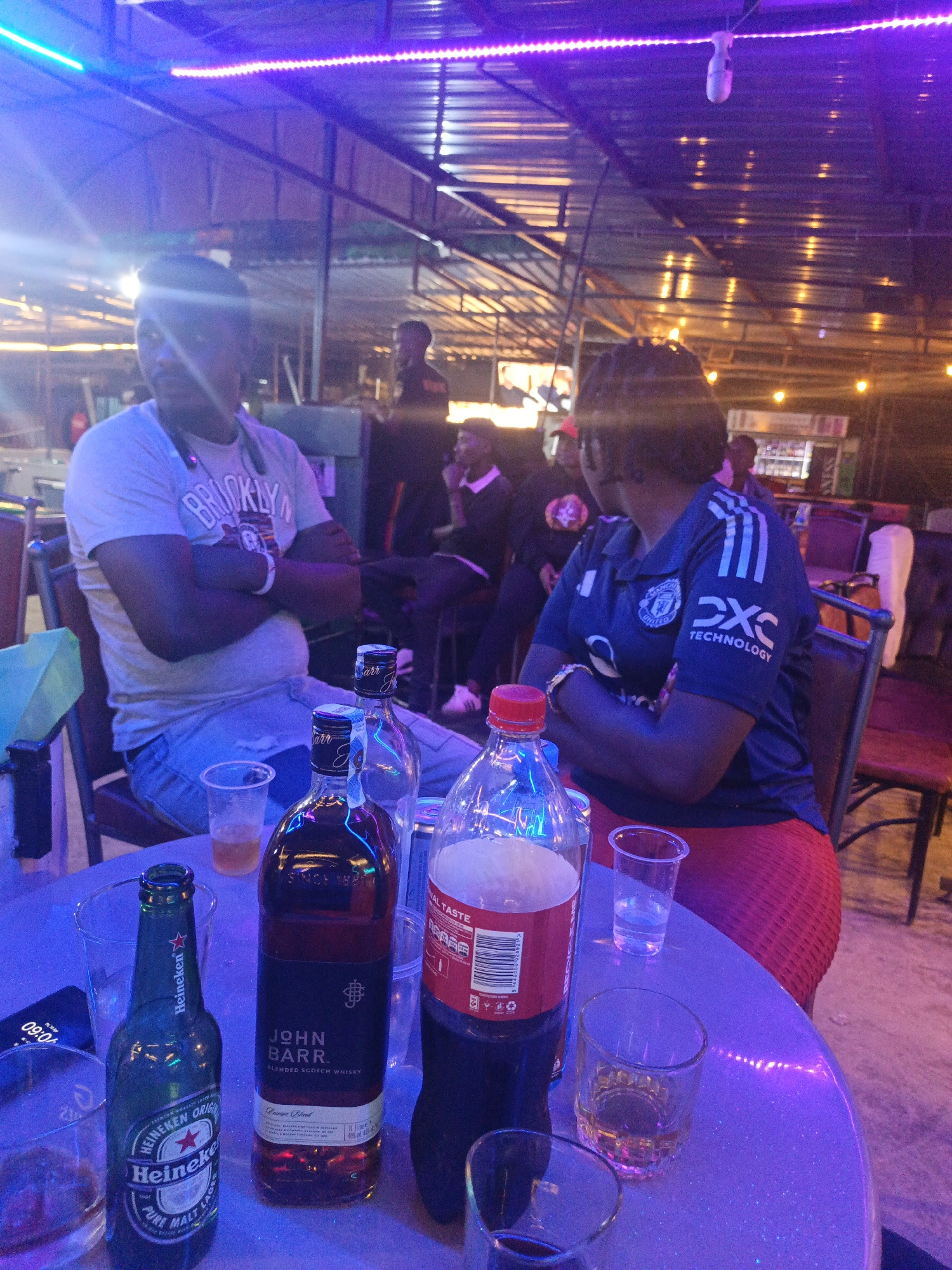 The image shows a table at an outdoor bar or restaurant, likely in Nairobi, Kenya, given the individuals present and the presence of local brands. Two people sit at the table: a man in a "Brooklyn" t-shirt and jeans, and a woman in a Manchester United jersey and red skirt. The table is littered with several bottles of alcoholic and non-alcoholic beverages including a Heineken beer, a bottle of John Barr blended scotch whisky, and a bottle of Coca-Cola. Several drinking glasses, some partially filled, are also visible on the table. In the background, other individuals can be seen seated, standing, or walking around, suggesting a moderately busy atmosphere. The environment is lit with colorful lights, including pink and blue lights strung above, indicating it is nighttime.