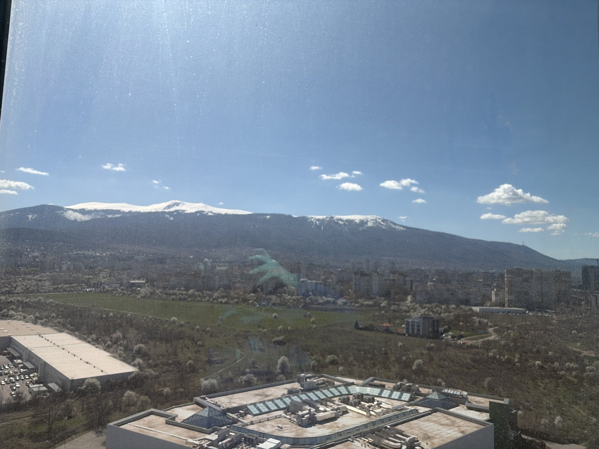 The image shows a view of Sofia, Bulgaria, looking towards Vitosha Mountain, from a high vantage point. The mountain has snow on its peaks. Below, the city is visible, with various buildings, greenery, and trees. In the foreground, the rooftop of a large building with skylights and various structures can be seen. A large warehouse building is to the left, with an adjacent parking lot full of cars. There is also a greenfield area between the warehouse, large building, and city.