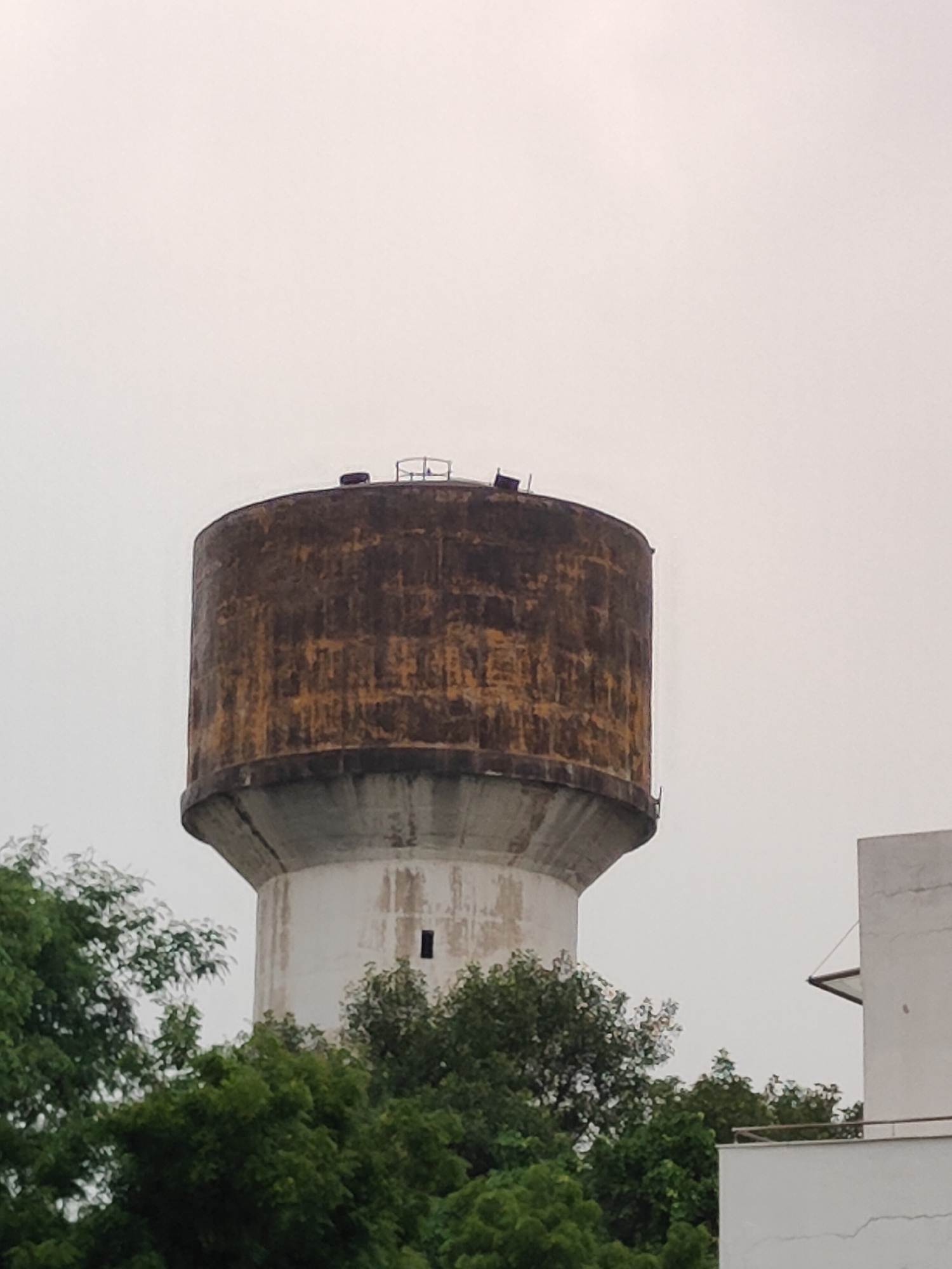 The image shows a tall, cylindrical water tower with a weathered, rusty-brown exterior on its upper half and a white, conical base. The tower stands against a pale, overcast sky. Lush green trees surround the base of the tower and extend into the foreground. To the right of the water tower, a portion of a modern white building with a triangular roof structure is visible. The scene appears to be set in an urban or suburban area, likely Gurugram (formerly Gurgaon), India, given the presence of trees and architectural styles common in the region. There are no people or visible actions taking place.