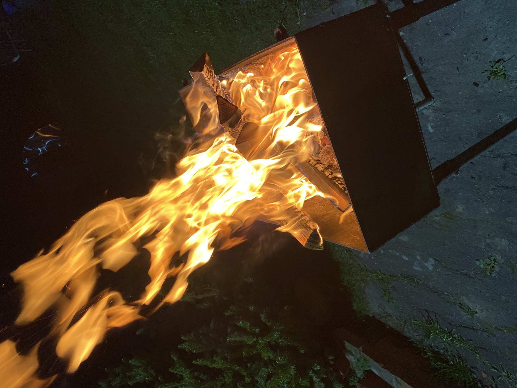 A metal fire pit is ablaze with flames consuming wooden logs in Красный Маяк, Russia. The fire pit stands on a concrete surface near some vegetation. The environment appears to be dimly lit, possibly at night.
