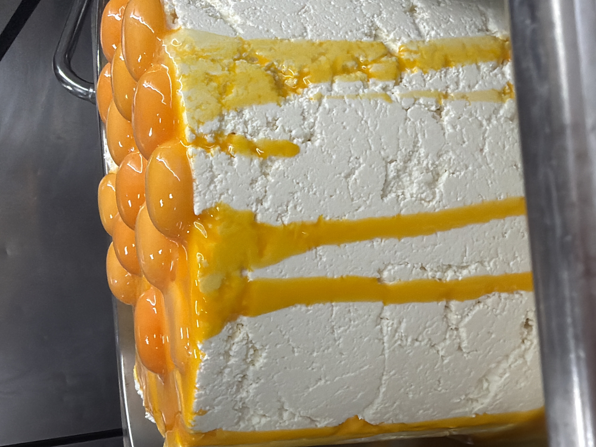 A close-up shot captures a rectangular dessert, seemingly in a stainless steel serving tray. The dessert has a layered appearance with a white, textured base, possibly consisting of soft cheese or a whipped cream-like substance. Drizzled or layered within the white base is a yellow, viscous substance resembling honey or a fruit compote. On the left side of the dessert, a vertical arrangement of small, glossy, orange fruit pieces (perhaps apricots or candied citrus) are aligned and glazed. The dessert is sitting in a metallic serving tray, and a stainless steel bar is visible on either side. The setting appears to be indoors, possibly a restaurant or bakery based in Belgrade, Serbia.